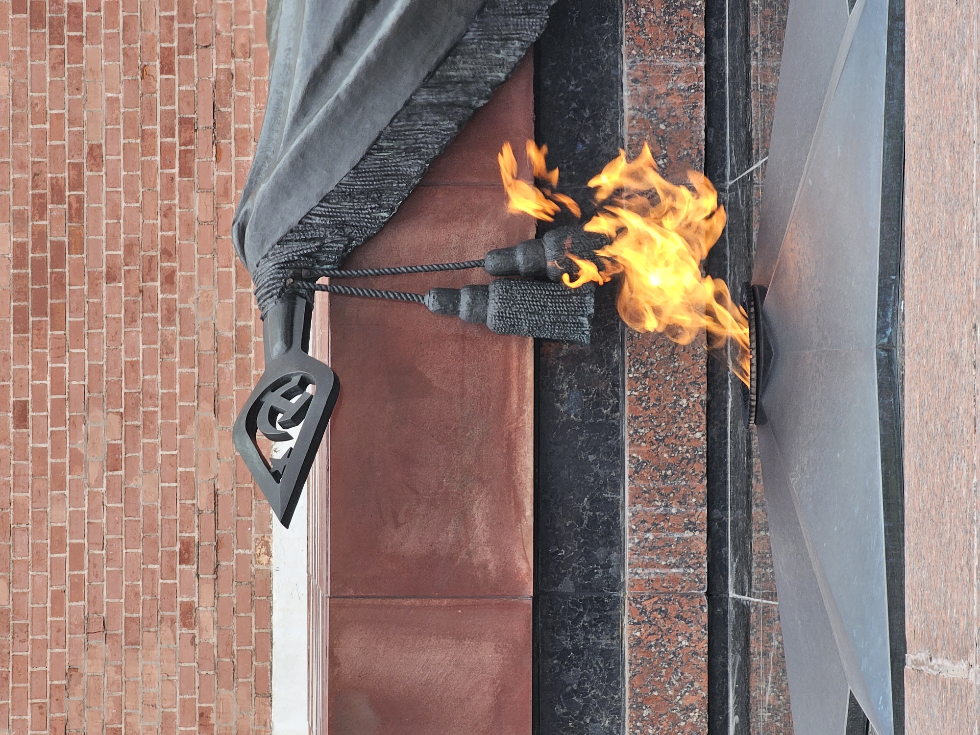 The image shows a close-up of a memorial site in an urban area. A perpetual flame burns in front of a stone structure, likely a monument or part of a larger memorial complex. The flame is bright and active, positioned centrally in the frame.
To the left of the flame, a grey drape or cover is partly visible, secured with a decorative tassel. The architectural elements surrounding the flame and drape are constructed from various materials, including brick and polished stone or granite. The textures and colors of these materials suggest a formal and solemn setting.
The background is predominantly architectural, with the brickwork providing a clear indication of the built environment. There is no visible signage to indicate the exact location, nor are there any people depicted in the image.