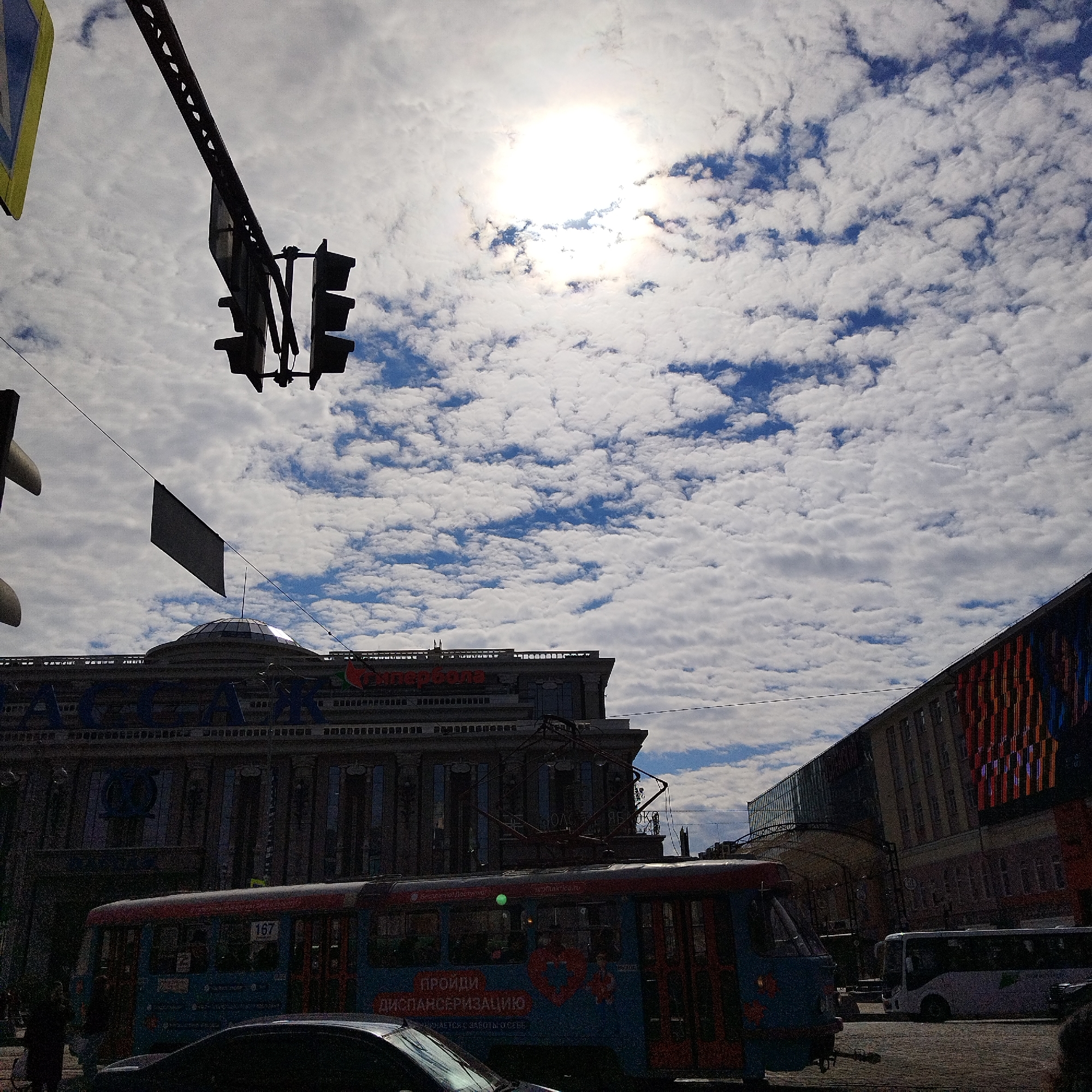 The image depicts a street scene in Yekaterinburg, Russia on a bright, sunny day with a sky filled with altocumulus clouds. A tram, light blue with red trim and marked with the number 167, is in the foreground. Behind the tram is a dark-colored car.
In the background, there is a building with the name "Passage" displayed prominently. To the right of the "Passage" building is a modern building with a colorful, vertically striped design on its facade. A white bus is parked near that building.
Overhead, there is a traffic light and a black flag hanging above the tram. There are also power lines or trolley wires visible. Pedestrians are present on the sidewalk near the tram.