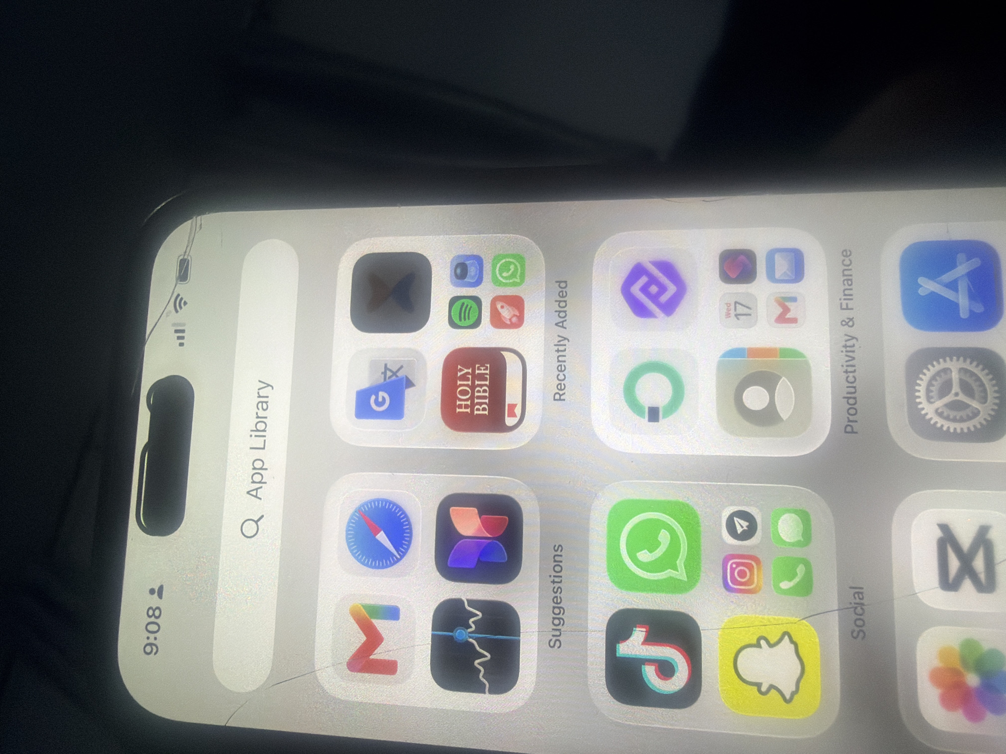 The image shows a close-up of a smartphone screen, displaying the app library. The time on the phone is 9:08. App icons visible include Gmail, Compass, Google, a graph, a butterfly, and the Holy Bible. Below these are categories like "Suggestions" and "Recently Added." Further down, social media apps like TikTok, WhatsApp, Snapchat, Instagram, and Telegram are visible, along with a category labeled "Social." The phone screen has visible cracks, particularly near the top and across the screen. The background is dark and out of focus. There is no indication of the city of Bukuru, Nigeria in the image.