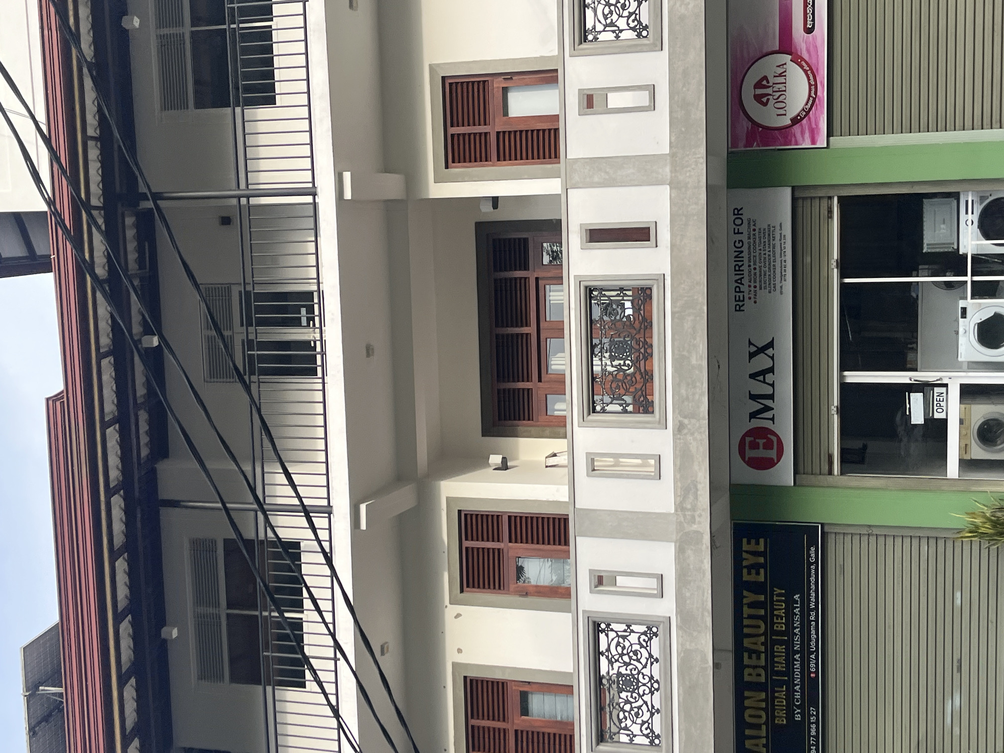 The image shows the exterior of a multi-story building with shops on the ground floor. The building has a white facade and a red tiled roof. There are several windows on the upper floors with wooden shutters. The ground floor features three storefronts. The leftmost store, labeled "SALON BEAUTY EYE," appears to be a beauty salon, offering bridal, hair, and beauty services. The middle store is an appliance repair shop, listing services for various electronics like TVs, audio systems, washing machines, fans, microwaves, ovens, toasters, blenders, and electric kettles. The rightmost storefront displays a pink logo with the name "LOSELKA." The shops are located at 691/A, Udugama Road, Walahanduwa, Galle, Sri Lanka. Two washing machines are visible through the open doorway of the middle shop. Power lines are strung across the upper portion of the building.