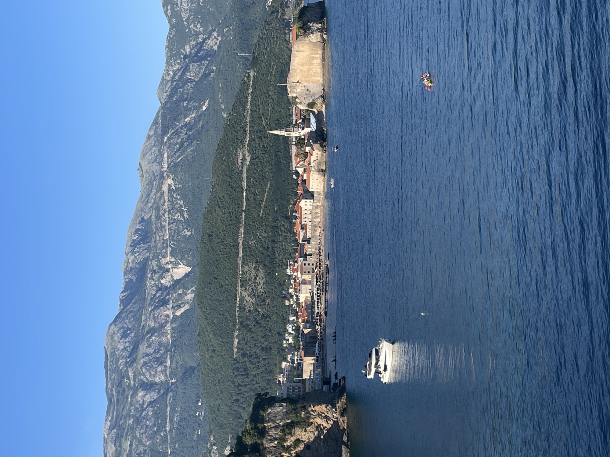The image shows Budva, Montenegro, with a mountain on the left, the city in the middle, and the Adriatic Sea on the right. A white yacht is near the shore. Further out in the water, a group of people are on an inflatable platform. Buildings with red tile roofs line the coast. The sky is clear and blue.