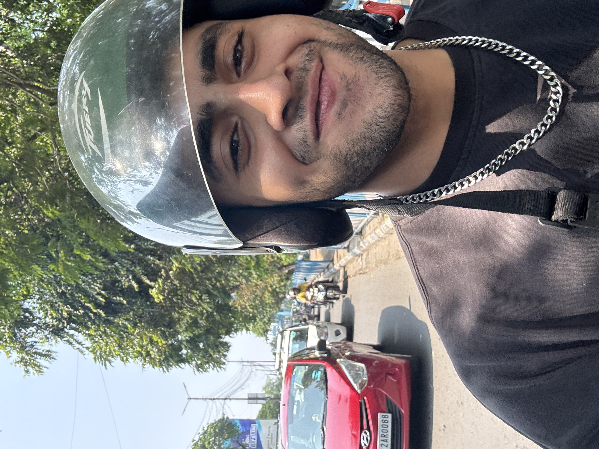 The image shows a man wearing a helmet and a silver chain, likely on a motorcycle, as he is positioned above a red car with the license plate "2AR0088." The backdrop shows a cityscape with trees, cars, motorcycles, and buildings. The signage visible in the background suggests the location is in Kolkata, India, indicated by Bengali script on the signs. There is traffic on the road, and the man seems to be pausing or moving slowly. The lighting suggests it is daytime.