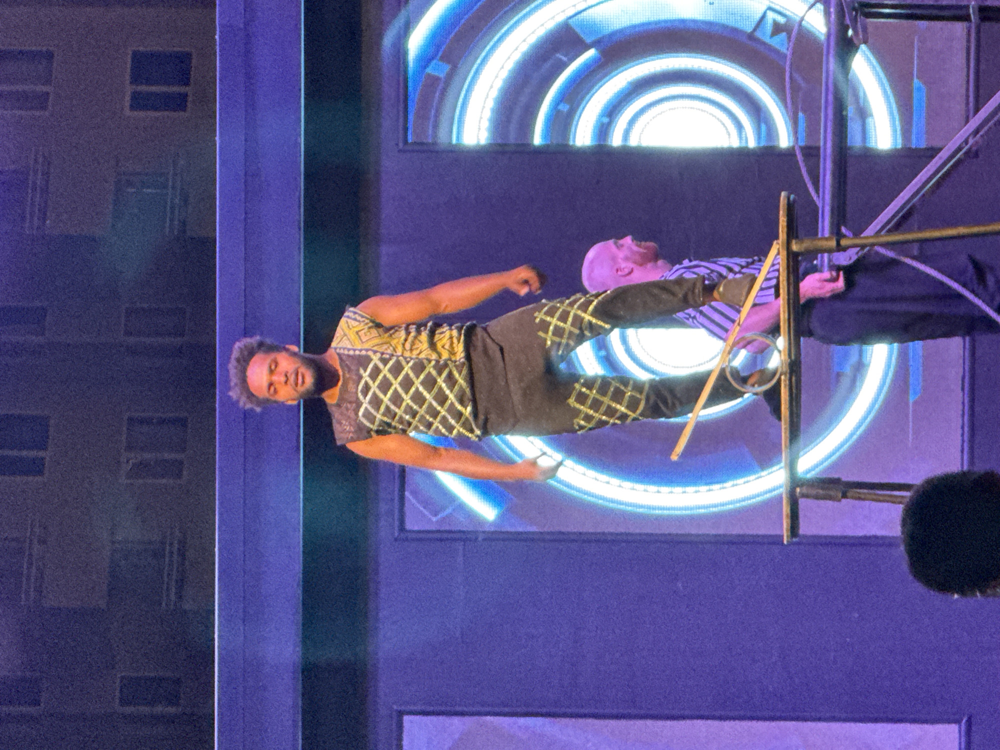 A man with dark, curly hair and a beard, wearing a sleeveless black and gold patterned top and black pants, stands on a narrow table on a stage. He is in the process of balancing a thin stick horizontally with his right hand, and another stick vertically with his left hand, which is bent at the elbow. The backdrop of the stage features large, illuminated blue and white circular patterns. To the right of the central figure, another man with a shaved head, wearing a striped shirt, is visible. The overall environment appears to be a performance or entertainment venue. The image is a photograph taken from a slightly low angle, capturing the performers from the chest up.