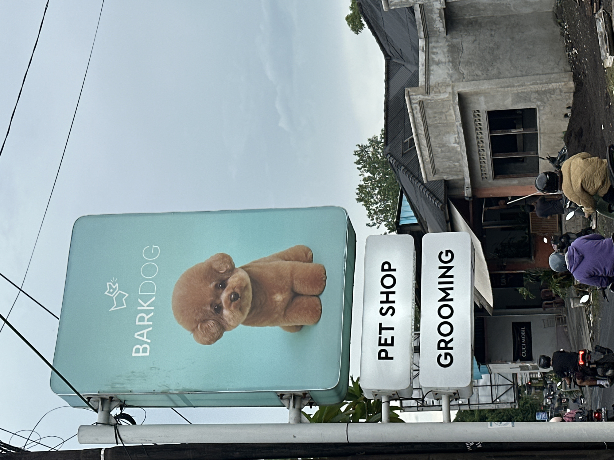 The image shows a "Barkdog" pet shop sign in Munggu, Indonesia. The sign is light blue with a picture of a brown poodle and the words "Barkdog", "Pet Shop", and "Grooming." The sign is attached to a white frame. In the background, there is a two-story building and several people on motorcycles. The sky is overcast.