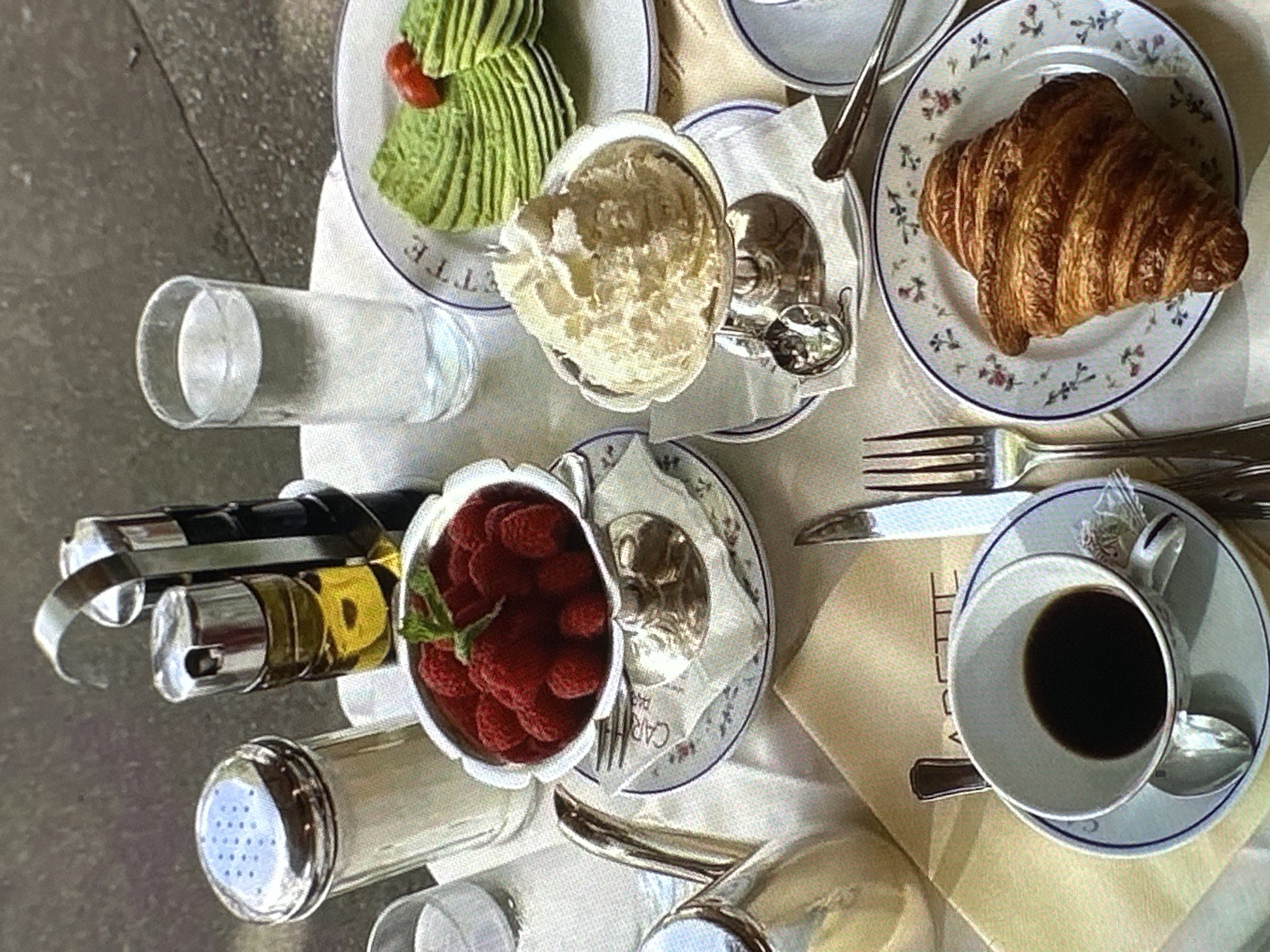 A still life photograph presents a table setting, likely in Yerevan, Armenia, composed of breakfast items. At the top left, a plate contains sliced avocado with a small red tomato. Adjacent to it, a silver container holds a fluffy, white, unidentified cream. A croissant rests on a decorative plate. Below, a clear glass filled with water is next to a condiment set containing a silver dispenser and oil and vinegar bottles. Fresh raspberries are served in a silver bowl. A cup of black coffee is placed next to a knife and fork.