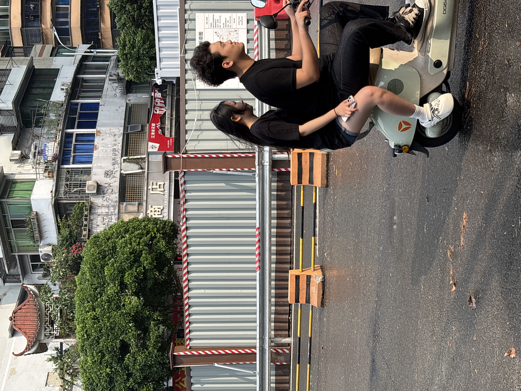 Two young people are on a small scooter on a street in Jiangjin, China. The rider is wearing a black t-shirt, black jeans, and black and white sneakers. The passenger is wearing a black t-shirt and denim shorts and is holding a phone. The scooter is light green and white. The background includes buildings, trees, construction barriers, and street markings. A sign with Chinese characters is visible on one of the buildings.