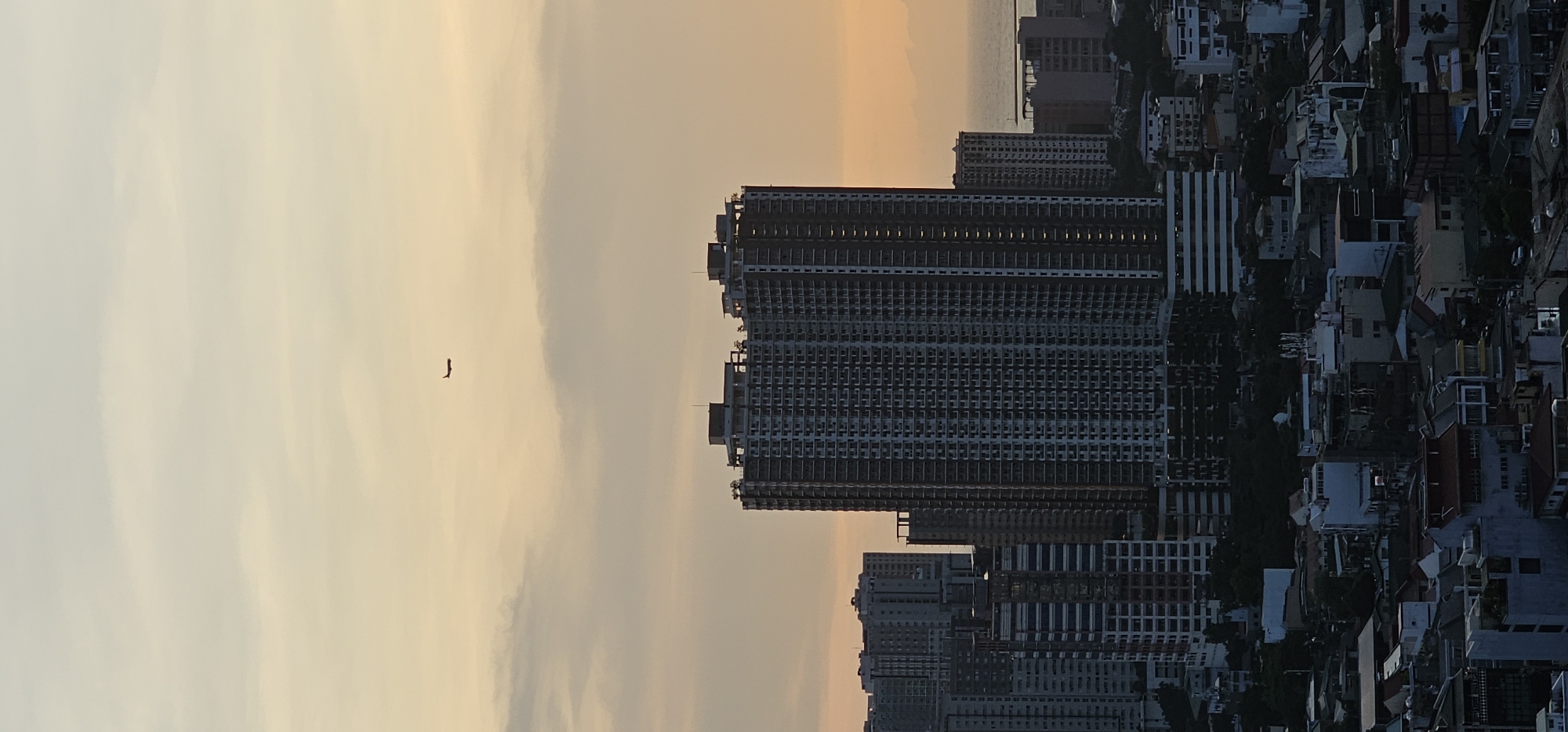 An aerial, vertical, wide shot captures a city skyline during twilight. The sky is a gradient of soft orange and pale yellow, with wispy clouds. A silhouette of an airplane is visible in the upper portion of the sky. Below, a dense urban landscape is dominated by numerous high-rise buildings. The most prominent structure is a very tall, modern building with a dark facade and many windows, centrally located. To the left and right of this central building are other skyscrapers of varying heights and architectural styles. The foreground shows a lower-density area with smaller buildings, rooftops, and glimpses of trees. The overall scene depicts a bustling city transitioning from day to night. The location is identified as Makati, Philippines.