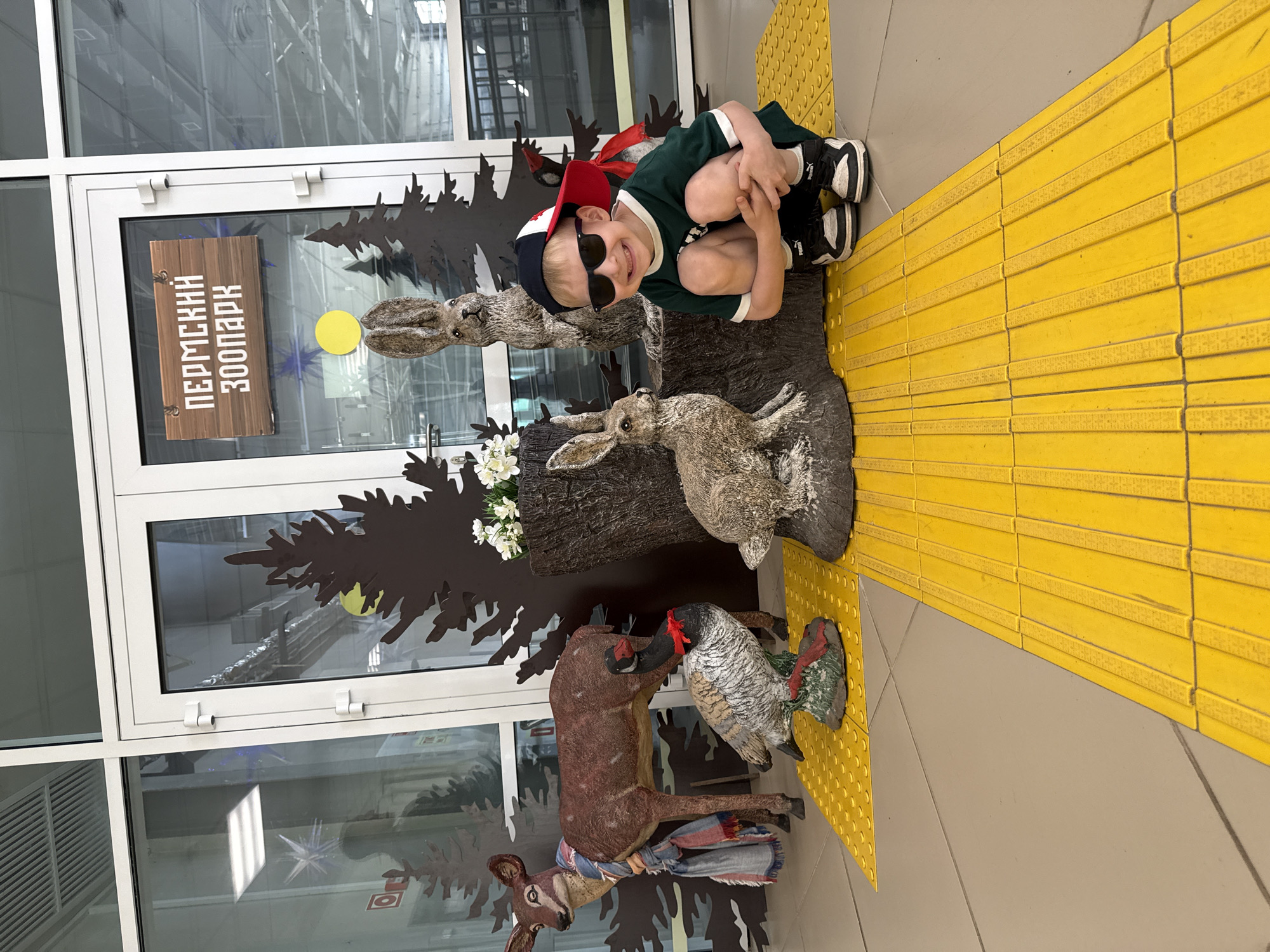 In a bright, indoor environment, likely the entrance of a building, a young boy is crouched down on a yellow tactile paving surface. He is wearing sunglasses, a green t-shirt, and a baseball cap. The boy is smiling at the camera. Behind him are several life-size animal sculptures: a deer wearing a scarf, a duck with a red bow around its neck, and two rabbits. A wooden sign with Cyrillic lettering, "ПЕРМСКИЙ ЗООПАРК," which translates to "Perm Zoo," hangs on a glass door in the background, indicating the location is the Perm Zoo in the city of Perm, Russia. The overall scene depicts a cheerful moment at the zoo entrance.