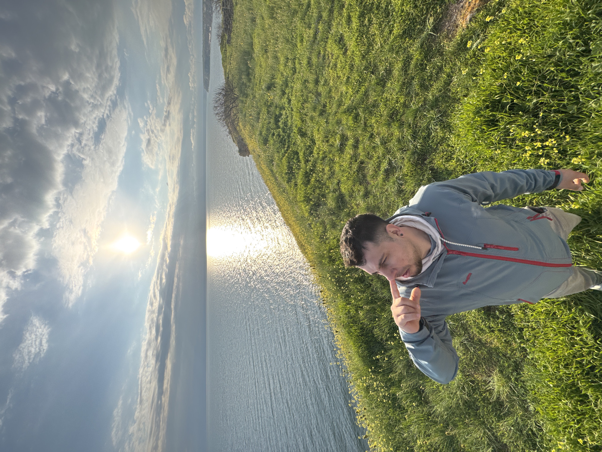 A person in a blue jacket and light pants is standing on a grassy hill overlooking a body of water under a partly cloudy sky. The sun is reflecting on the water's surface. The location is likely Balgarevo, Bulgaria. The person is pointing at his nose.