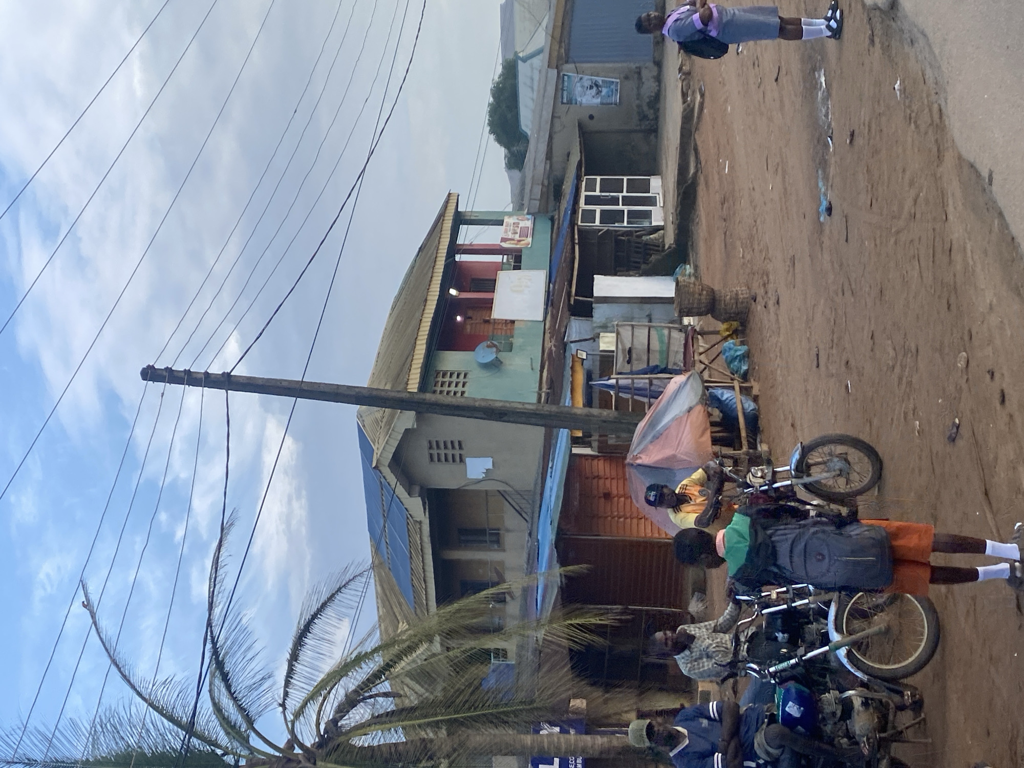 A group of people are gathered around motorcycles on a dirt road in Ikorodu, Nigeria. A young person in a school uniform, consisting of a grey shirt, grey shorts, white socks, and black and white shoes, stands on the road facing away from the camera. They are wearing a black backpack. Several motorcycles are parked, with some people seated on them. One person wearing a yellow shirt and a black baseball cap is leaning against a motorcycle. Another person, wearing a plaid shirt and glasses, is seated on a motorcycle. The scene appears to be outdoors, with buildings visible in the background. A palm tree is also visible, with its fronds reaching towards a cloudy sky. Electrical wires crisscross the sky. The ground is a mixture of dirt and sand. The lighting suggests it is daytime, possibly late afternoon, given the cloud cover. No specific emotion is evident on the faces of the individuals. Some signs are visible on the buildings, but the text is not legible.
