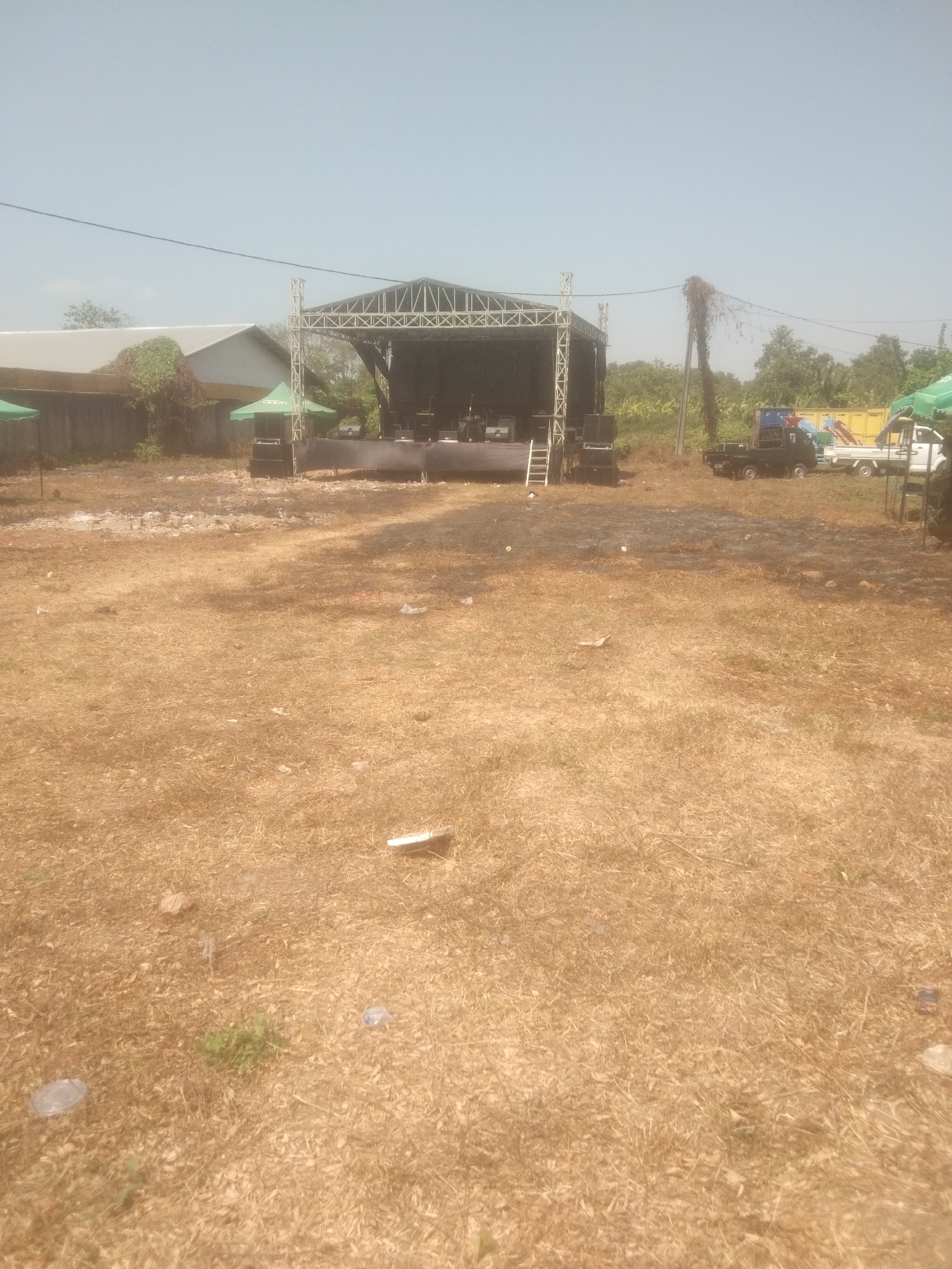 The image depicts an outdoor event setup in what appears to be a rural or semi-rural area in Kecamatan Haurgeulis, Indonesia. A stage structure with a metal truss roof stands center-frame, equipped with sound equipment and lighting. Several large speakers are positioned on either side of the stage. In front of the stage, the ground is covered with dry, brown grass and some scattered debris, including plastic cups and wrappers, suggesting it might be a temporary event space or a recently used area.
To the left of the stage, a building with a light-colored roof is visible, partially obscured by vegetation. Several green tents are set up near the building and the stage. To the right of the stage, a black pickup truck and a white truck are parked, along with a larger yellow truck further in the background. A utility pole with vines growing on it stands near the trucks. The sky above is clear and blue. The overall scene suggests preparations or a wind-down period for an outdoor performance or gathering.