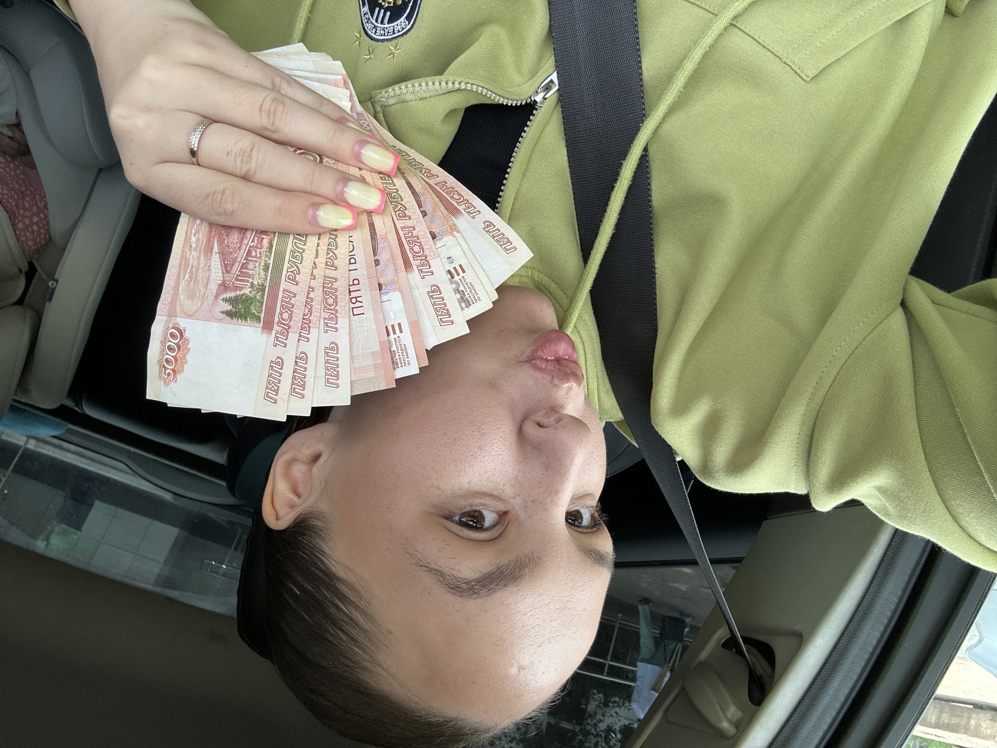 A young woman with her hair pulled back is sitting in the driver's seat of a car, holding a fan of Russian 5,000 ruble banknotes in her right hand. She is wearing a lime green hooded sweatshirt. Her lips are pursed. Her fingernails are painted with a French manicure featuring yellow tips and pink accents. A silver ring with small clear stones is on her right ring finger. The seatbelt is visible across her chest. In the background, through the car window, a person in a light blue shirt and green pants is walking away, pulling a dark rolling suitcase. The setting is the city of Chaikovsky, Russia, likely on a sunny day.