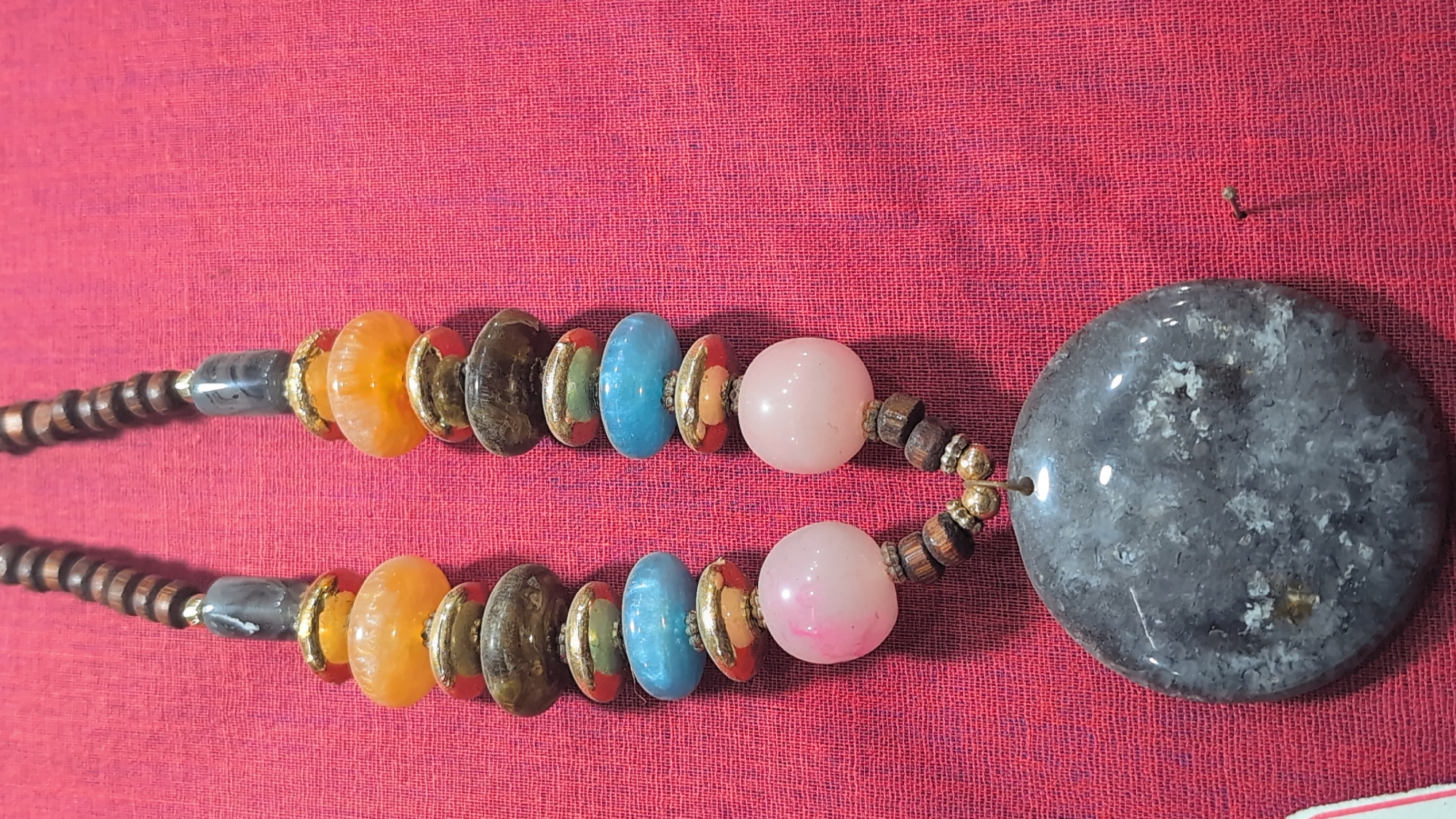 A close-up, overhead shot shows a beaded necklace on a red textured fabric. The necklace has two strands of beads that come together at a large, flat, oval-shaped pendant. The pendant is a marbled grey stone. Above the pendant, both strands are adorned with wooden beads and smaller, round pink beads. Further up, the strands feature alternating blue and orange beads, separated by gold-colored, fluted spacers and dark, oval-shaped beads. The top of the necklace is made of dark brown wooden beads.