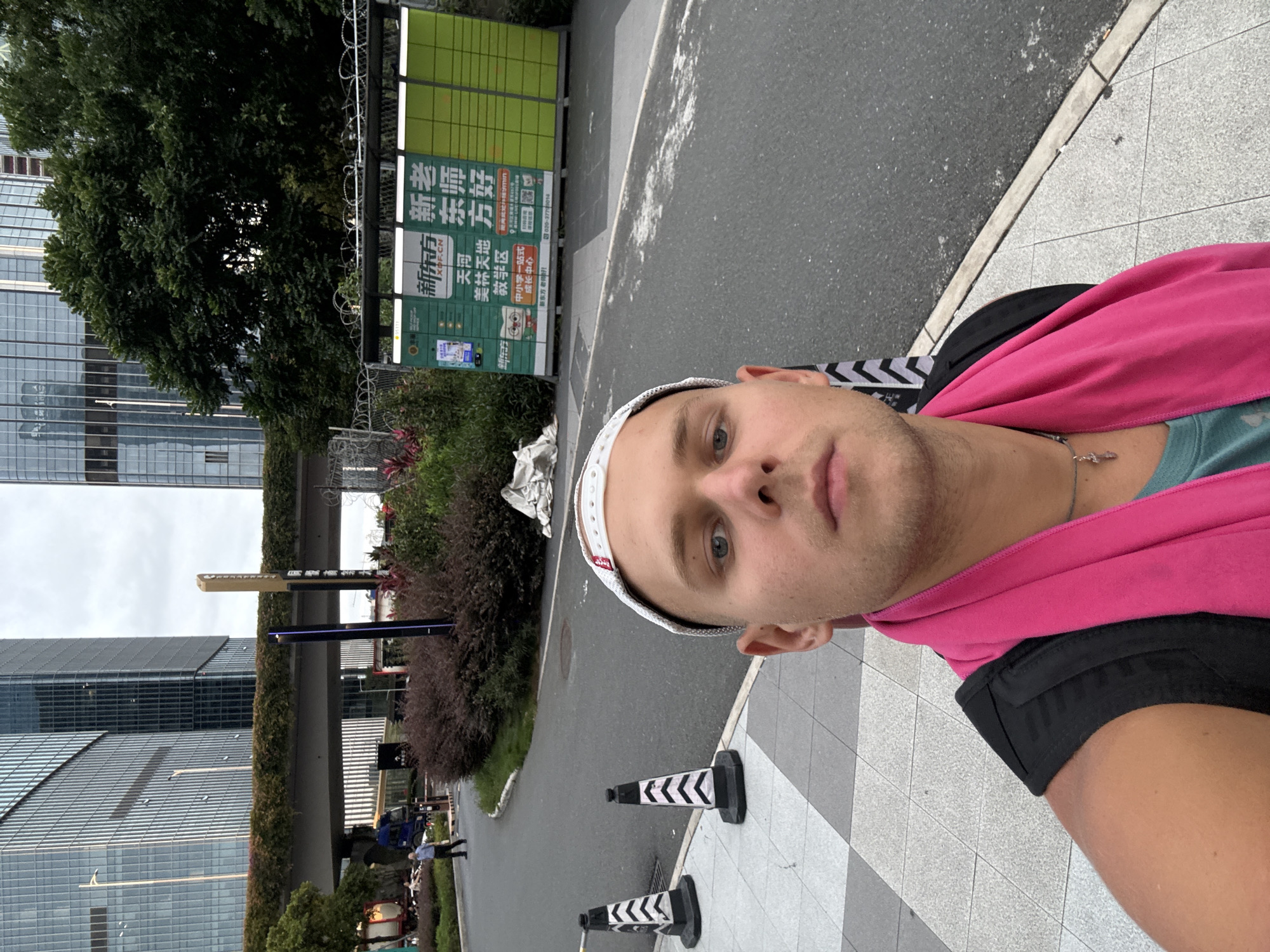 A male is taking a selfie outdoors in a modern urban environment, likely in Huangpu District, China. He is wearing a white baseball cap and a bright pink scarf draped around his neck. In the background, there are tall, modern glass buildings, a elevated walkway covered in greenery, and a street with traffic cones. To the right, a green and yellow automated parcel locker system is visible, with Chinese characters displayed on its panels. The man is standing on a paved walkway with a checkered pattern. The overall scene suggests a commercial or business district.