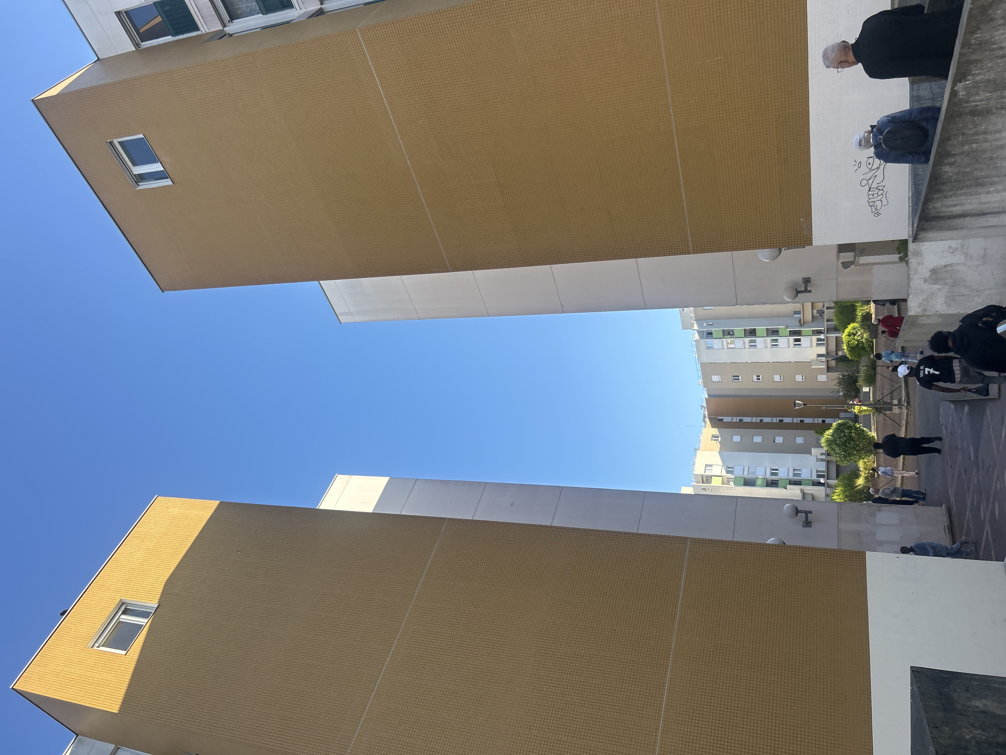 This upward-looking, portrait-oriented image captures a dense urban environment under a clear blue sky. Two prominent, multi-story buildings, characterized by their striking golden-brown tiled facades, flank the central void, with their lower sections featuring white or light grey cladding. Rectangular windows with white frames are visible on both structures.
In the mid-ground and background, further apartment buildings are visible, appearing in shades of grey, white, and some with green panels, typical of modern residential complexes. A clear, bright blue sky fills the space between the structures, indicating a sunny day.
Several people are gathered on what appears to be an elevated walkway or courtyard area to the right side of the frame. In the upper right, an older man with grey hair, dressed in a dark coat, stands alongside a person in a blue jacket and white cap leaning against a white wall. This wall features black graffiti. Further down the walkway, other individuals are present, including one person wearing a black top with "VINI JR" visible in white text on the back, and another in a red outfit. The people appear to be socializing or passing through the area.
Notable details include outdoor wall lights on the mid-ground buildings and some scattered greenery like trees and bushes. The strong sunlight casts shadows, suggesting midday or early afternoon. The architecture and density of the buildings are consistent with the provided location context of Noisy-le-Grand, France, a commune known for its residential developments.