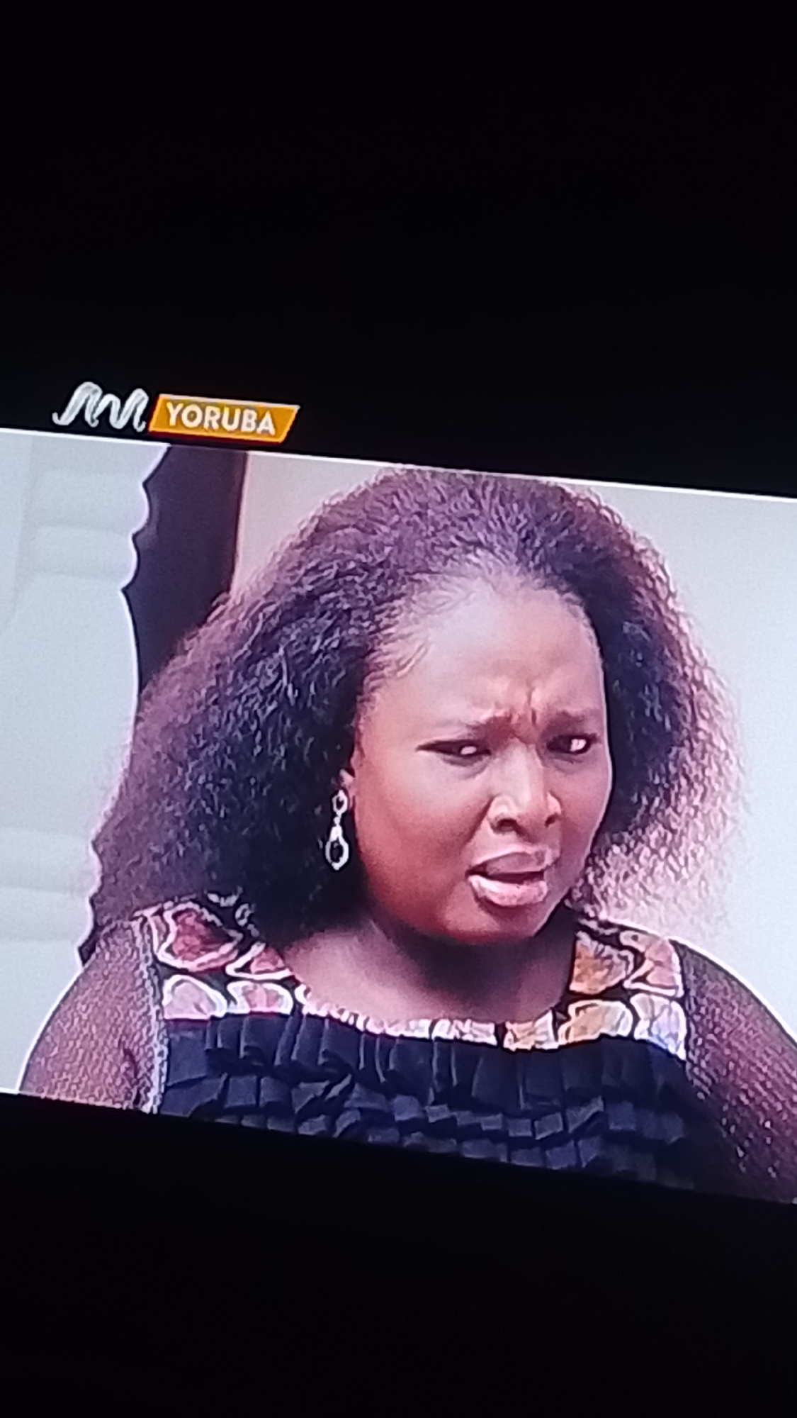 A medium shot shows a woman from the chest up, looking distressed or angry. She has dark, curly hair and is wearing a black dress with a patterned neckline. She has a dangling earring on her left ear. A logo in the upper left corner of the screen reads "M YORUBA." The background is indistinct, but appears to be an indoor setting. There is no indication of the city of Lokoja, Nigeria, in this image.