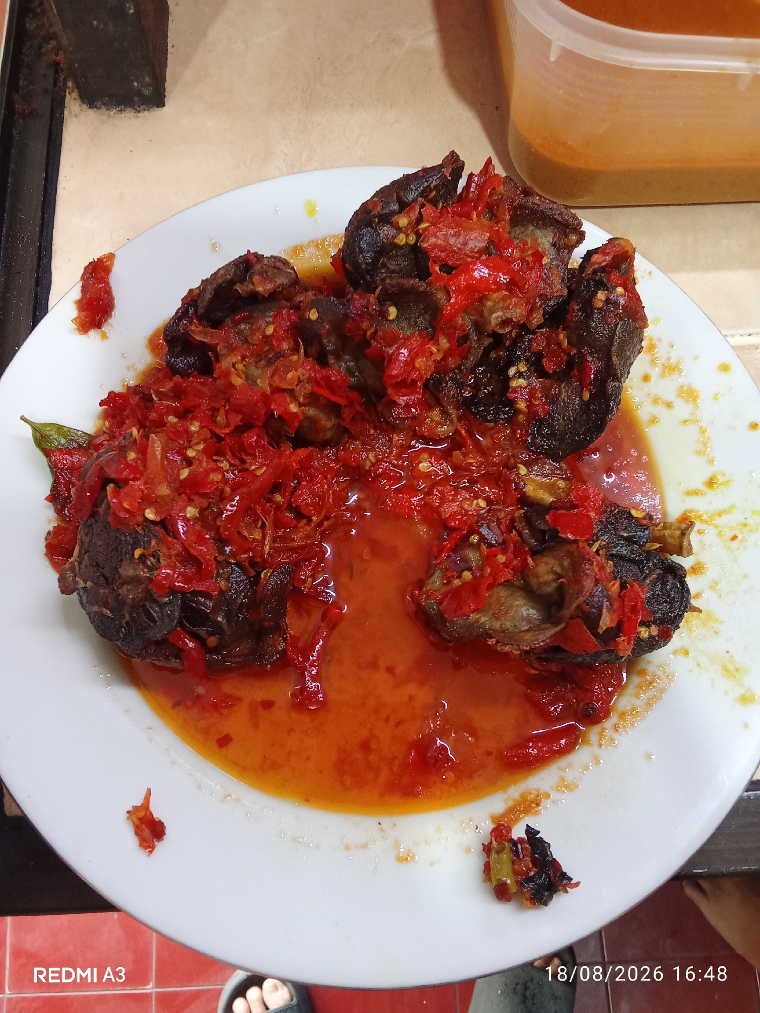 In Banaran, Indonesia, a close-up, top-down shot captures a white plate filled with a dish of dark, braised meat covered in a vibrant red chili sauce. The meat appears to be in chunks and has a slightly charred texture. The chili sauce is chunky, with visible pieces of chili pepper, some of which are whole or roughly chopped, and appears to be quite oily. The plate is placed on a beige, speckled surface, possibly a countertop, with a portion of a black, textured object visible in the upper left corner. To the right of the plate, a partially visible translucent plastic container holds a thick, orangey-brown sauce. The lower portion of the image shows a red tiled floor, with a black sandal and a bare foot entering the frame from the bottom right. The bottom left of the image displays the text "REDMI A3" and the bottom right corner shows the date and time "18/08/2026 16:48".