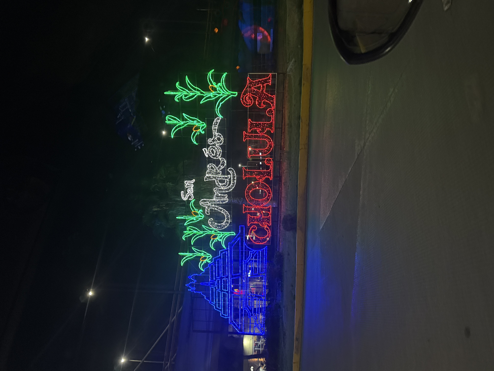 The image depicts a nighttime view in San Andrés Cholula, Mexico. An illuminated sign spelling "San Andrés Cholula" is displayed. The word "San Andrés" is in white lights, above the word "Cholula" written vertically in red lights. Above the name, there are three lit green palm tree symbols. Below the name is a blue-light silhouette of a building. The sign sits on a median between two lanes of a road that is lit up by street lights. A vehicle side mirror is visible in the upper right corner.