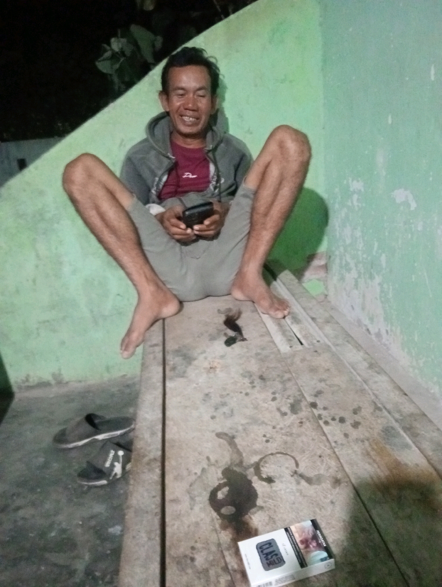 A man with a gray hooded jacket over a maroon t-shirt sits on a wooden surface with his legs spread. He is holding a dark-colored mobile phone in his hands. The background consists of a light green wall with some debris. On the floor to the left of the wooden surface, a pair of black sandals are visible. On the wooden surface, there are several dark stains and a pack of "Class Mild" cigarettes. The lighting suggests an indoor environment at night. The presence of the "Class Mild" cigarette pack indicates this scene likely took place in Indonesia, as this is a popular brand there. Without further information, it is impossible to definitively pinpoint the exact location within Tanjung Pandan.