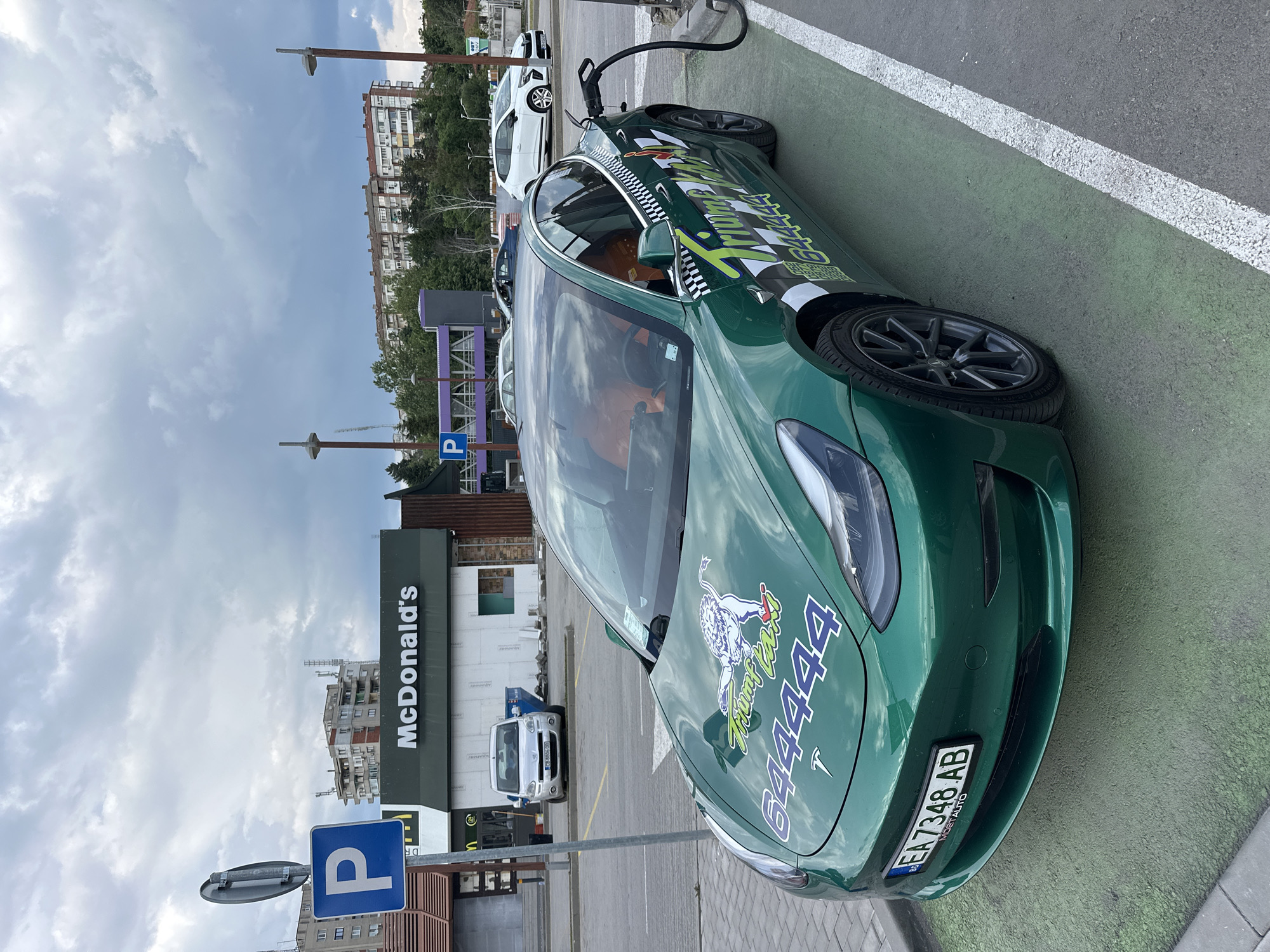 A green Tesla car with a distinctive paint job is plugged into a charging station. The vehicle has license plate "EA7348 AB" and is parked outside a McDonald's in Veliko Tarnovo, Bulgaria. Another vehicle is parked nearby. The sky is cloudy.