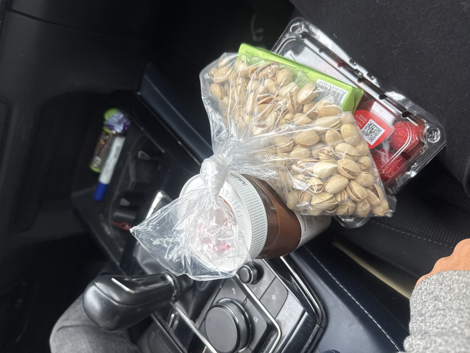 The scene depicts the interior of a vehicle in Mexico City, Mexico, observed from a slightly elevated perspective, likely from the passenger seat or the lap of an occupant. A collection of packaged food items is centrally positioned, resting on or against a person's leg, which is visible on the right side of the frame, clad in dark pants.
The primary objects include a clear plastic bag, tied at the top, filled with in-shell pistachios, situated prominently. Beneath this bag, a green rectangular package is partially visible, bearing the text "100%" and "Producto de México." Below these, a clear plastic clamshell container holds fresh red strawberries, featuring a red and white label with a QR code. To the left of the pistachio bag, a large jar containing a dark brown substance, consistent with Nutella, is present with a white screw-top lid. Resting on top of this jar is another clear plastic bag, also tied, containing a reddish liquid or fruit substance.
In the midground and background, components of the vehicle's interior are visible. To the left, an automatic transmission gear selector with a black knob is present, adjacent to a control panel featuring silver-accented rotary knobs and buttons. Further into the dashboard, a storage cubby contains a blue marker pen, a white marker pen, and a crumpled candy wrapper with purple and green coloring. A person's arm, covered by a grey sleeve, is visible on the far left of the image.