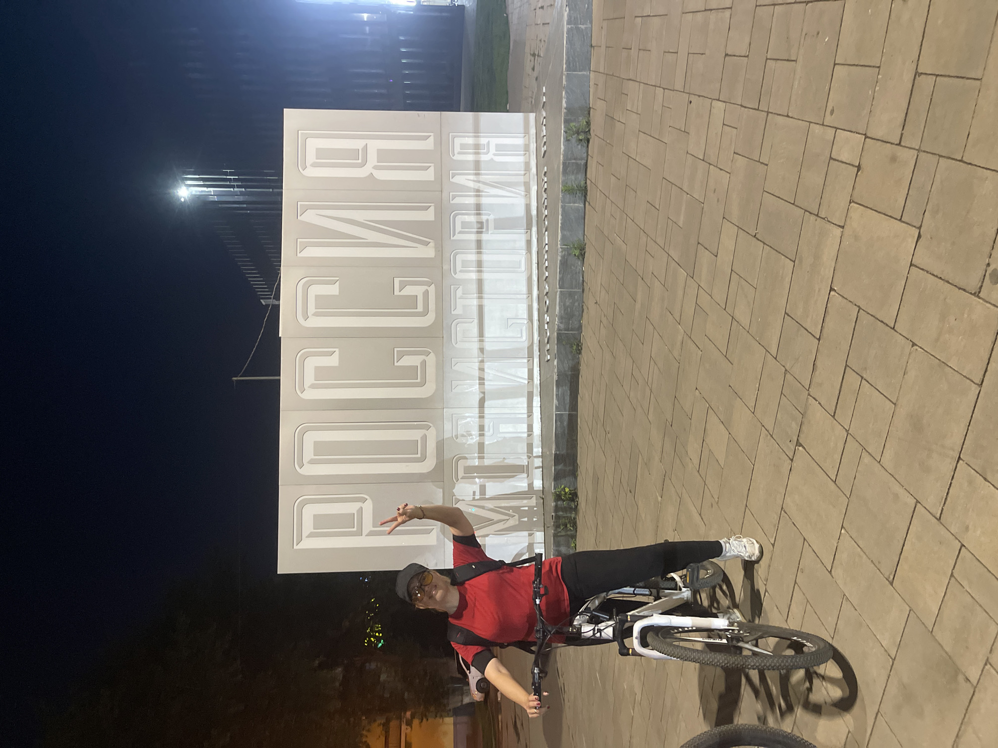 A woman is posing for a photo on a bicycle in front of a large white sign that reads "РОССИЯ ИСТОРИЯ" (Russia History) at night in Novokuybyshevsk, Russia. The woman is wearing a red t-shirt, black pants, a black baseball cap, and sunglasses. She is holding up two fingers in a peace sign. The sign is illuminated by lights, and the background is dark with some trees and buildings visible. The ground is paved with square tiles.