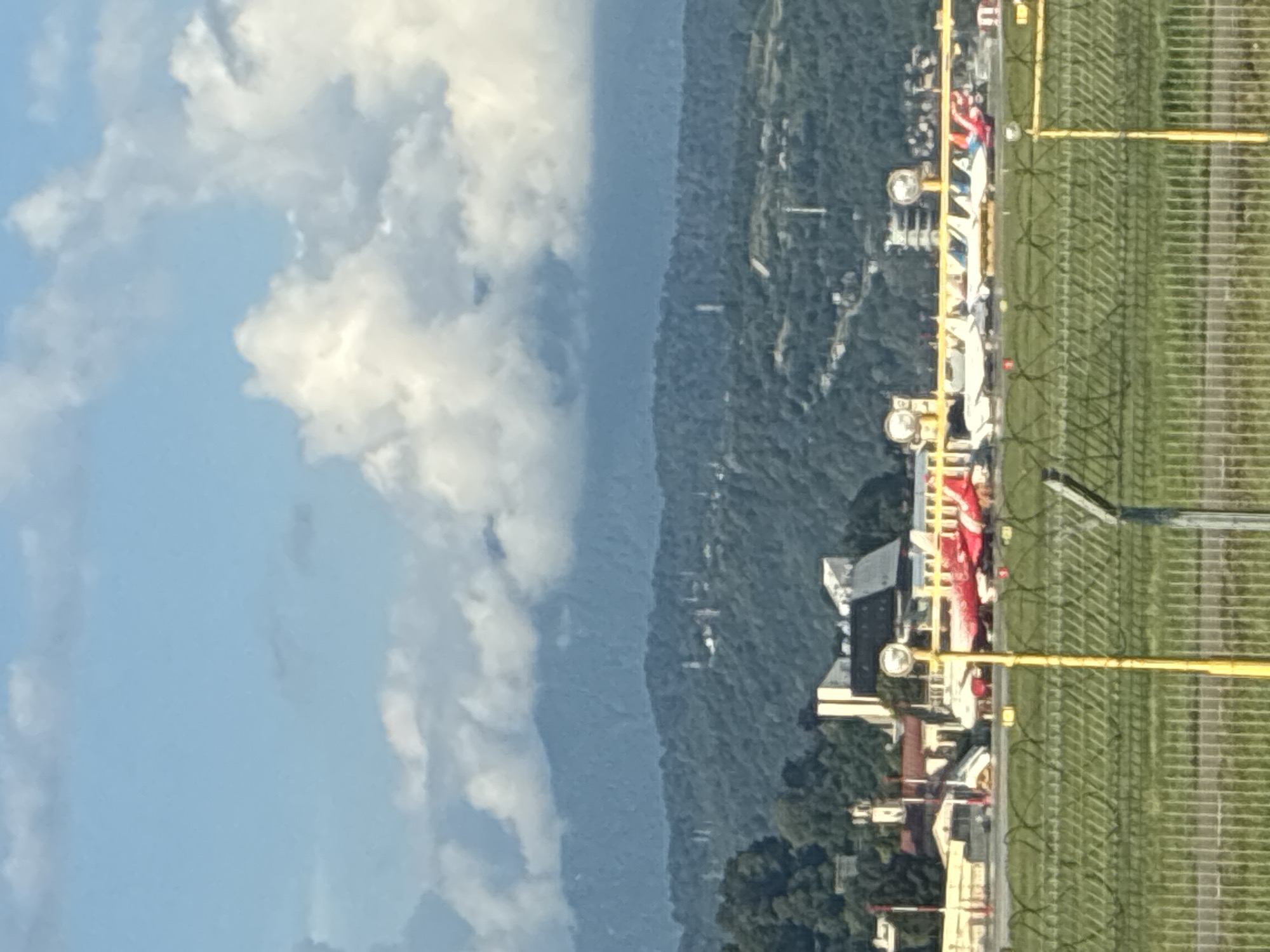 The image captures a view of Sochi International Airport (AER) on a clear day, with lush green hills and mountains forming a backdrop. Several airplanes, predominantly white with red accents, are parked on the tarmac. A chain-link fence with barbed wire runs across the foreground, separating the viewer from the airport operational area. Buildings, possibly airport terminals or support structures, are visible behind the fence. The sky is a brilliant blue, dotted with fluffy white clouds, and the distant mountains are shrouded in a soft, hazy blue. The overall scene suggests a typical day at a coastal airport nestled in a mountainous region.