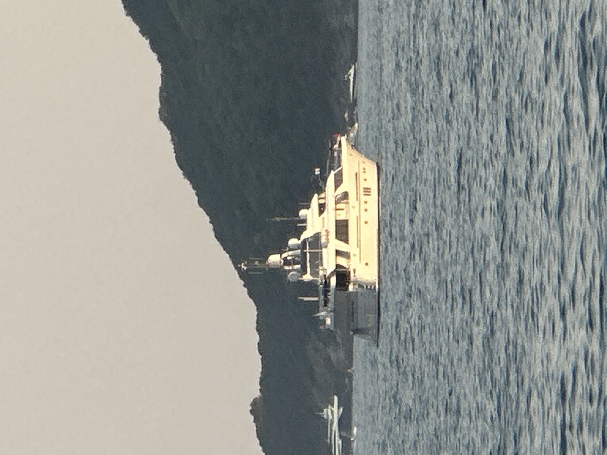 An outdoor, slightly hazy, daylight image shows a wide shot of a large, white motor yacht with multiple decks and windows on the water. The yacht is the primary focus, positioned slightly to the right of the frame. Behind the yacht, a dark green, forested hill rises from the water, forming the background. The sky above is a pale, hazy beige. In the distance, to the left of the main yacht, another, smaller boat with a white hull and dark superstructure is visible. Further to the right and further in the distance, a very small, dark boat is visible on the water. The foreground is filled with choppy, dark blue water with visible ripples. The scene appears to be a coastal area, possibly in or near Budva, Montenegro, given the characteristic topography. No people are clearly visible on the yacht or in the water.