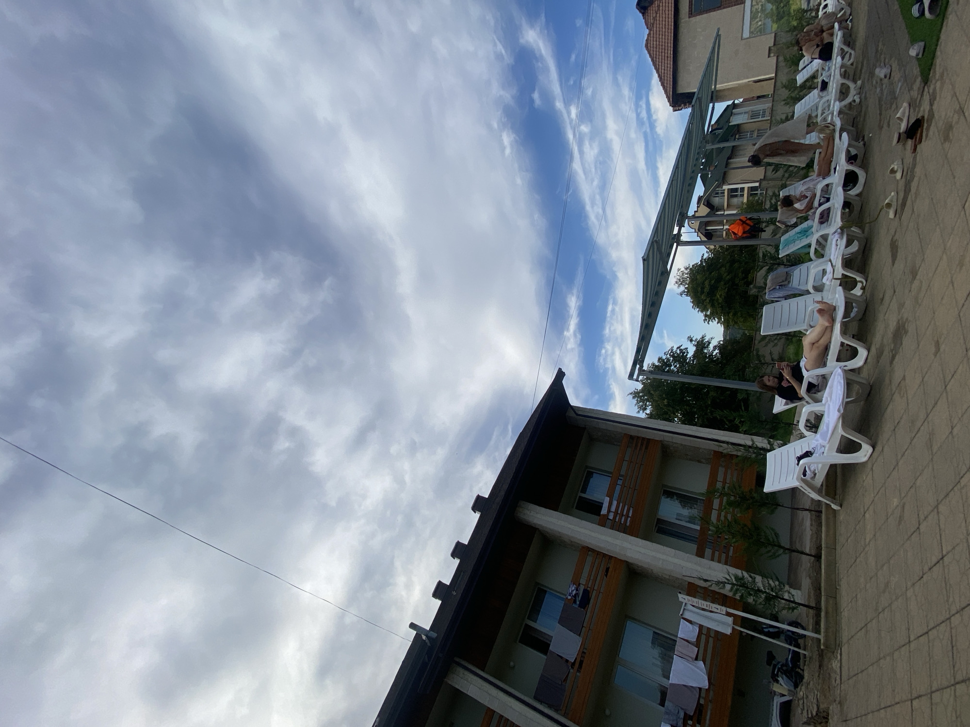 In the city of Molovata Nouă, Moldova, a low-angle shot captures a resort setting with a building and lounge chairs under a cloudy sky. The building, a multi-story structure with balconies, is on the left. Clothes hang from the balconies. In the foreground, a paved area hosts a row of white lounge chairs, several occupied by individuals sunbathing or relaxing. Some chairs are covered with white towels. Further back, a covered canopy provides shade for more lounge chairs. People are seen interacting casually, some looking at their phones, others in conversation. A life vest is visible on the canopy structure. The overall scene depicts a tranquil day at a resort.