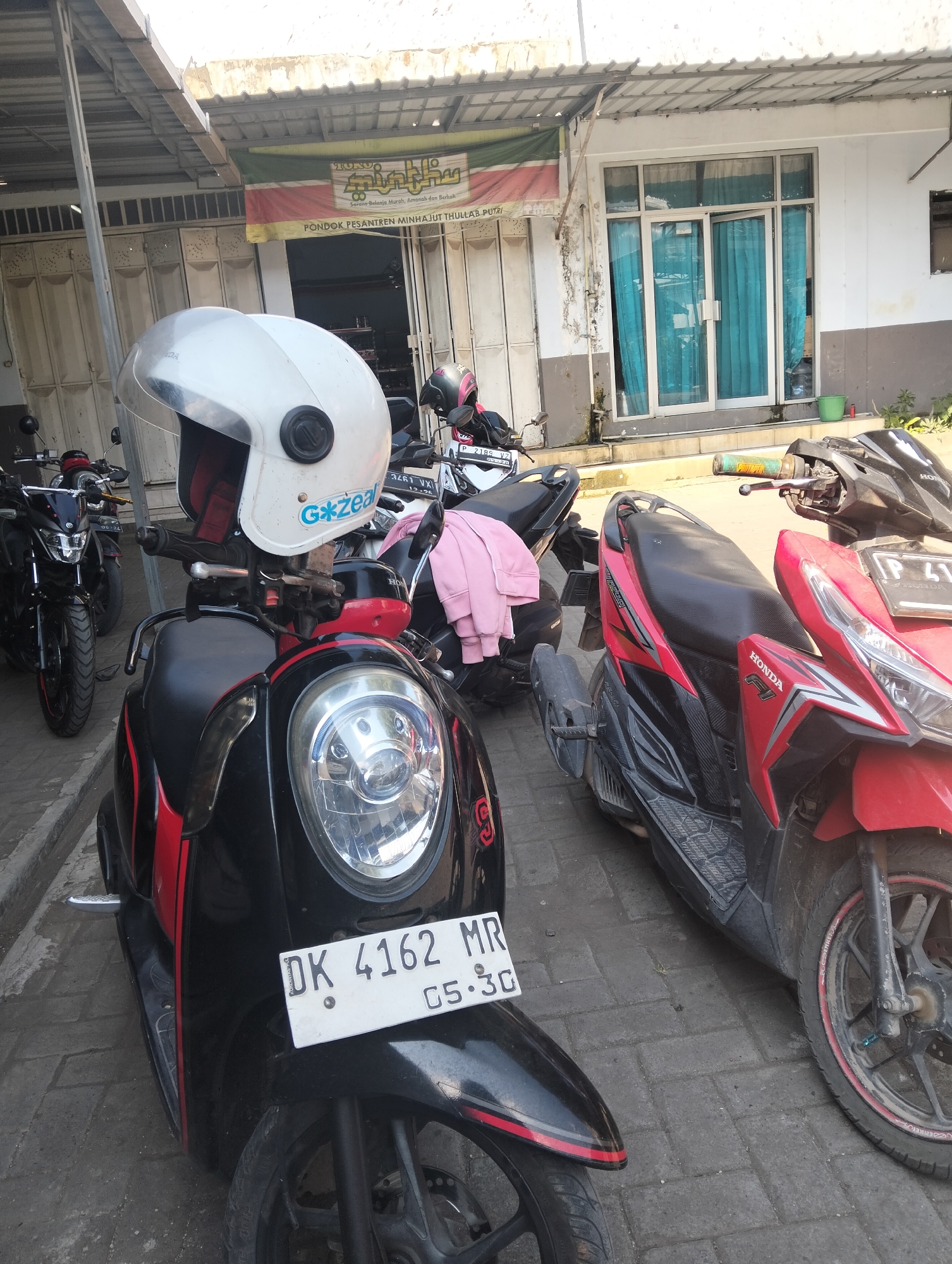The image captures a street scene in front of a building with a metal awning, likely in the city of Wringin Putih, Indonesia, as indicated by a banner that reads "Pondok Pesantren Minhajut Thullab Putri". Several motorcycles are parked, their fronts facing the viewer.
The closest motorcycle is black and red, with a white helmet placed on its handlebars. The helmet has a blue and white sticker that reads "G*zeal". A license plate on this motorcycle reads "DK 4162 MR 05.30".
To the right of this motorcycle is a red and black Honda motorcycle. Further to the left, another black motorcycle is visible, its front wheel prominent in the frame. Behind these, more motorcycles are parked, some with their handlebars turned.
The building in the background has large glass doors with teal curtains visible behind them. The ground is paved with interlocking bricks. The overall impression is of a sunny day with a focus on parked vehicles in an urban setting.