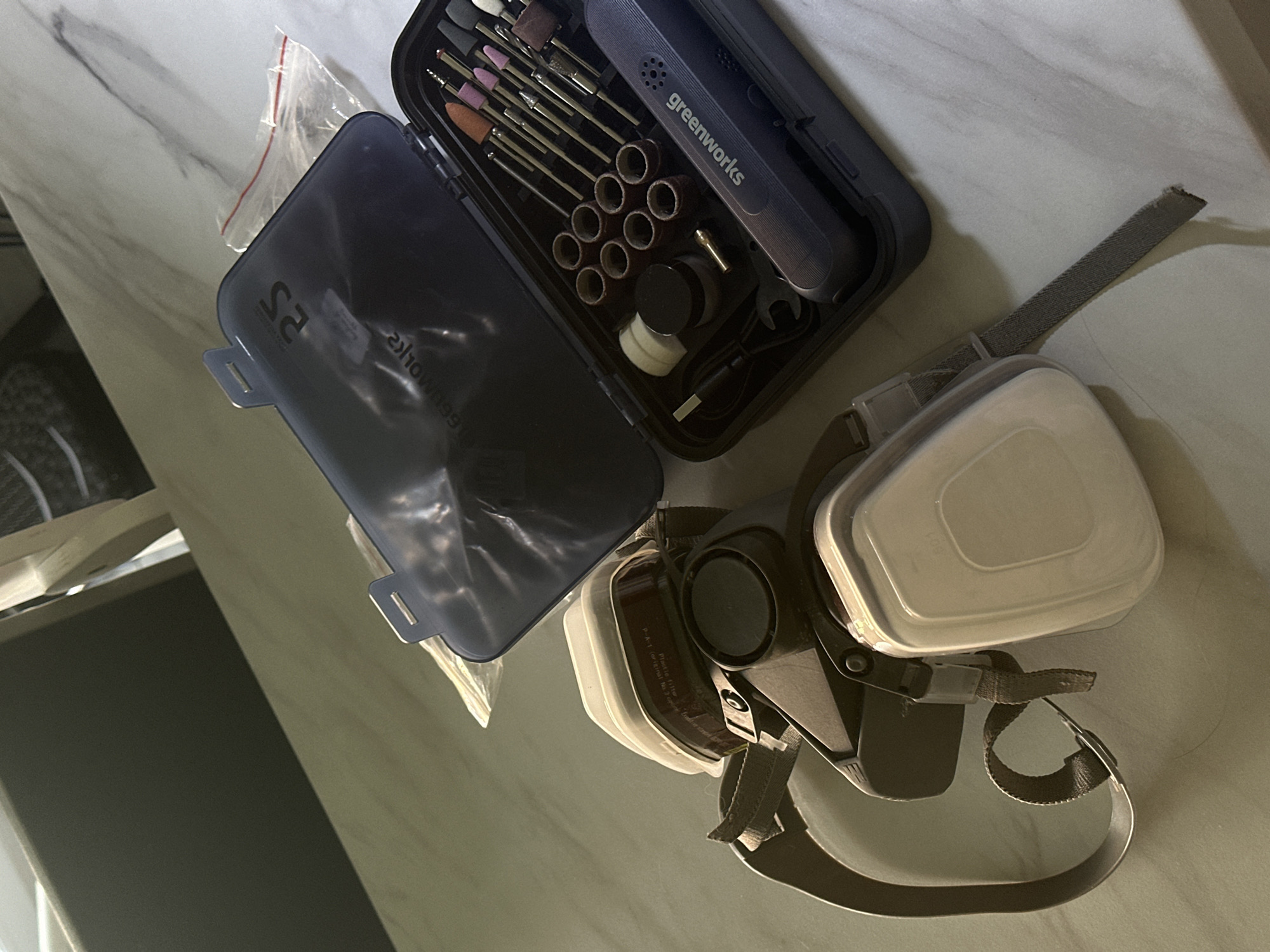 An overhead, tilted shot displays a rotary tool kit and a respirator mask laid out on a light-colored, possibly marbled, surface in an indoor setting in Kuda, Russia. The lighting appears to be artificial.
The primary objects are an open, dark blue or black case containing a rotary tool and its accessories, and a multi-part respirator mask. The rotary tool, branded "greenworks," is visible inside its organized case, surrounded by various attachments including grinding stones, sanding drums, and other bits. A small wrench, a charging cable, and what appears to be a small jar of polishing compound or similar accessory are also neatly arranged within the case. Reflective text inside the case lid shows "2S" and what appears to be a reversed "greenworks" logo.
Adjacent to the tool kit is a respirator mask, positioned with its filter cartridges facing upwards. The mask is dark grey and light beige, featuring two large, rectangular filter cartridges attached to the main body. Visible text on one of the cartridges includes "Plastic filter," "P-1," "for industrial air supplies only," and instructions like "original No, replace," and "Replace filters regularly." The mask has several adjustable straps, one of which extends across the frame. A small, clear plastic bag with a red zip-lock is situated between the tool case and the respirator.
The arrangement suggests the items are either recently used, being prepared for a task involving fine detail work, or stored. No people are visible, and no specific activity is taking place.