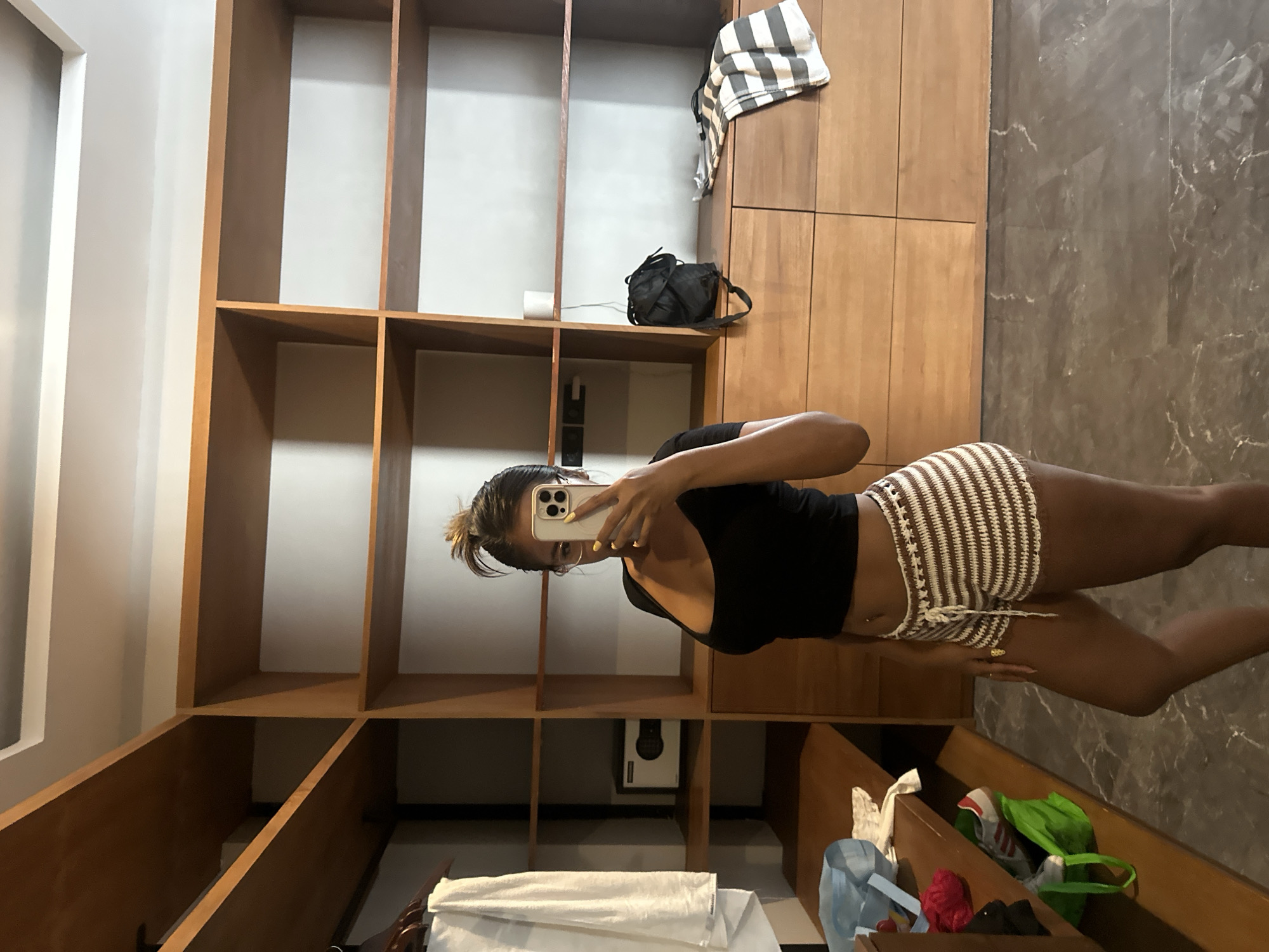 A woman is taking a mirror selfie in a room with a large wooden closet in North Kuta, Indonesia. She is wearing a black crop top and striped crocheted shorts. The closet has several open shelves and drawers, some of which contain bags, clothes, and a safe. The floor is tiled with a dark marble pattern.