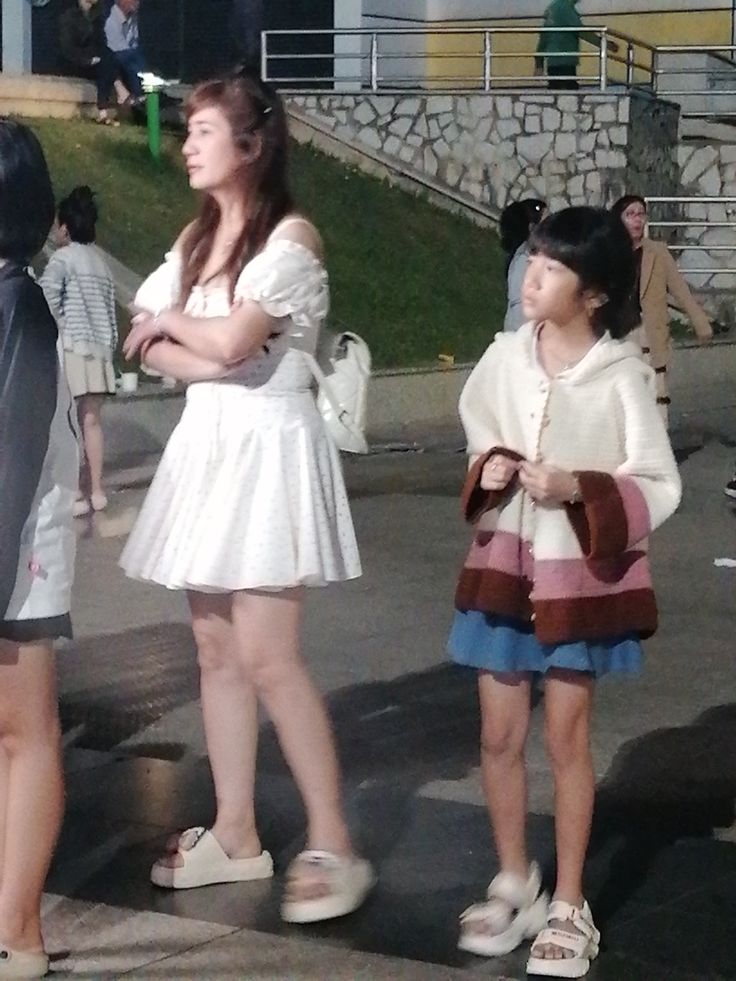 A medium-shot photograph shows two young women and other people in the background, outdoors in what appears to be a public space at dusk. The woman in the foreground on the left is wearing a white off-the-shoulder dress with puff sleeves and a floral pattern. Her arms are crossed, and she is looking to her left. She has long, brown hair and is wearing white platform sandals. To her right, a younger girl is wearing a cream-colored hooded cardigan with horizontal stripes of brown, pink, and white, over a blue skirt. She is also wearing white platform sandals and is holding onto the front of her cardigan. Her hair is dark and cut in a bob. In the background, other individuals are visible, including a person in a green jacket on a raised walkway and others seated on steps. The setting appears to be a park or a pedestrian area with paved ground and grassy slopes. The lighting suggests it is evening. One of the visible sandals on the younger girl has text on the strap, but it is not legible. The overall impression is a casual outing in a lively urban or recreational area.