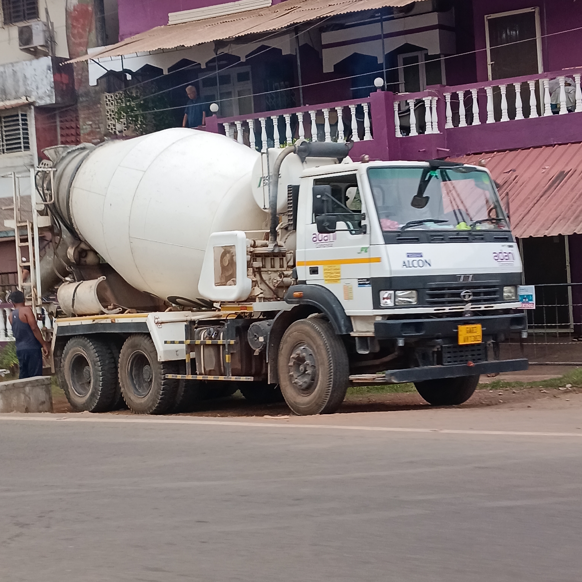 A parked white concrete mixer truck with "ADANI CONCRETE" and "ALCON" markings is located on the roadside in Sangolda, India. The truck has six wheels and a ladder attached to the mixing drum. A man wearing a tank top stands near the rear of the truck. In the background is a two-story building with purple exterior walls and a white balustrade on the second-floor balcony. A man stands on the balcony.