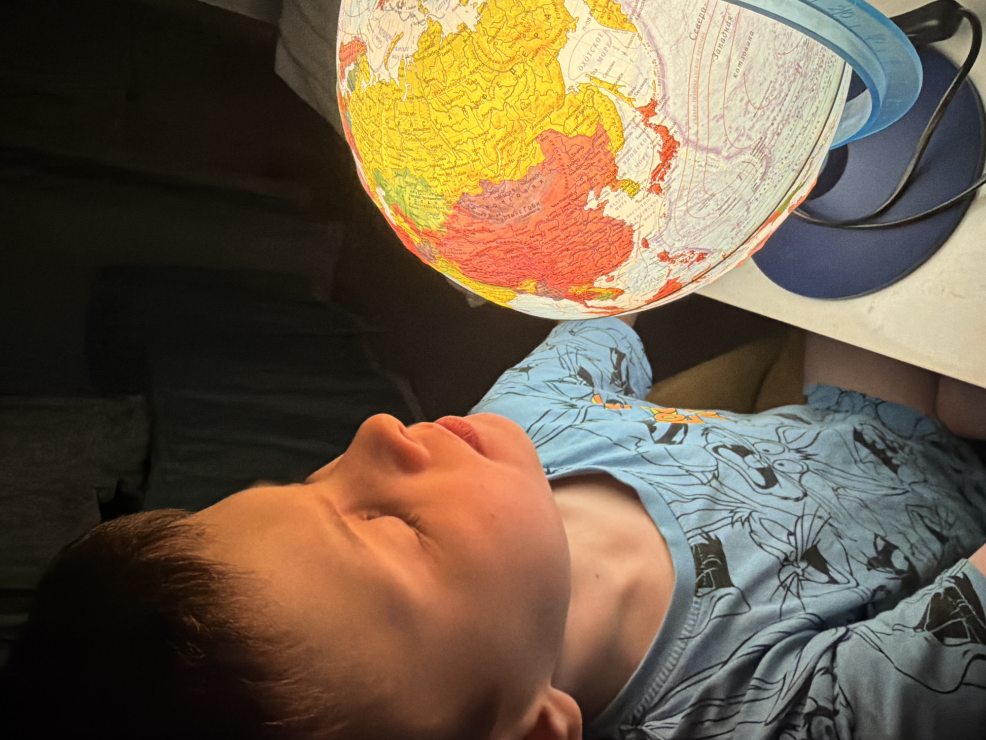 A boy lies on his back, eyes closed, wearing a light blue t-shirt with cartoon graphics. Above him is a globe illuminated from within, showing a map of Asia. The globe is mounted on a blue stand on a white surface. The background is dark and out of focus, suggesting a dimly lit room. The location is not discernible from the provided image.