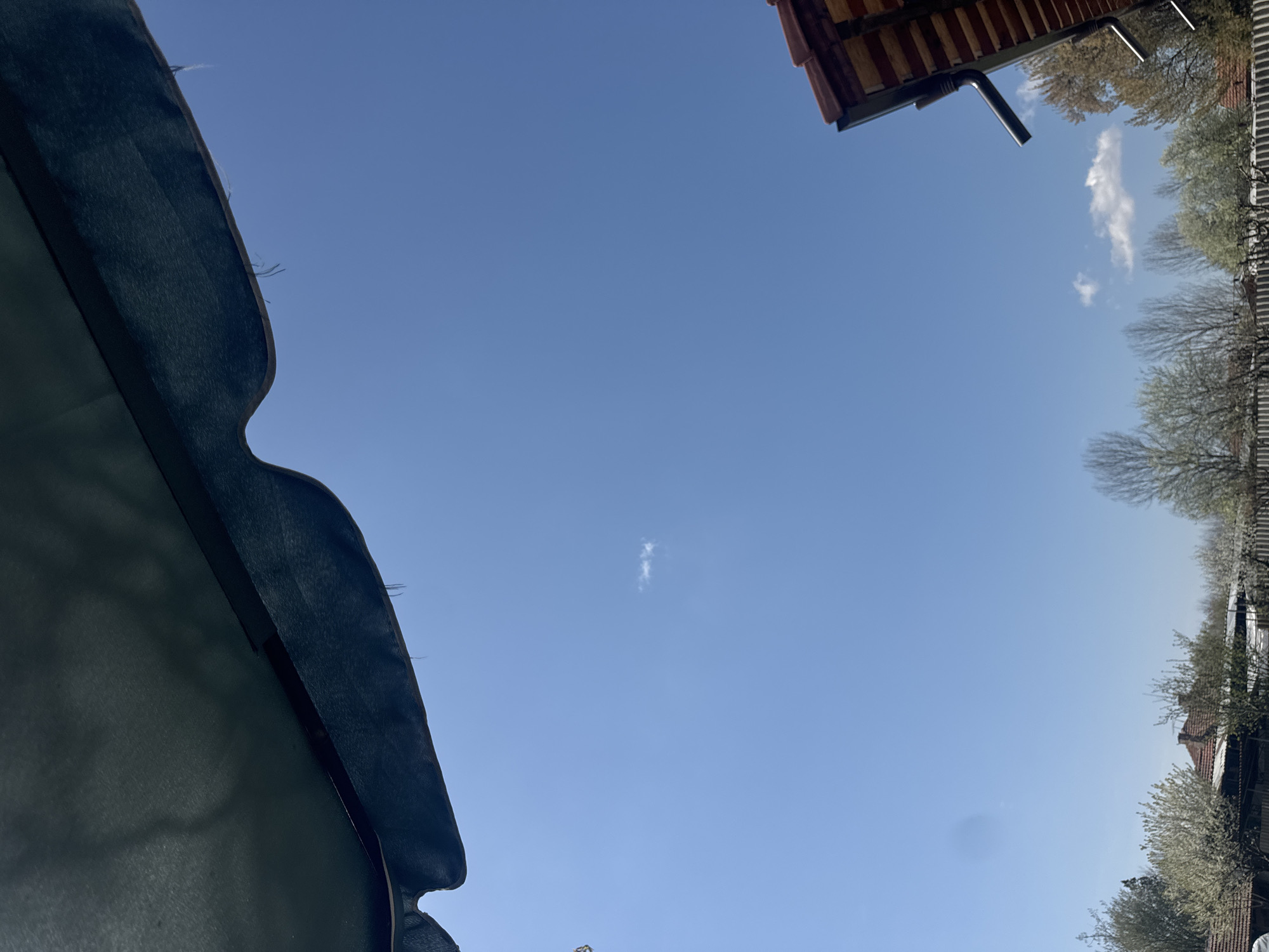 The image depicts a view looking upwards. The sky is predominantly blue with a few wisps of clouds. In the lower-left corner, there is a dark, possibly green, fabric or awning-like structure. In the upper-right corner, there is a roof with orange tiles and a dark drain pipe. Along the right side of the image, there are trees with budding leaves and the edges of buildings. The scene is likely an outdoor space, possibly a backyard or garden. I am unable to confirm the location is in the city of Lehchevo, Bulgaria based solely on the image. There are no people visible.
