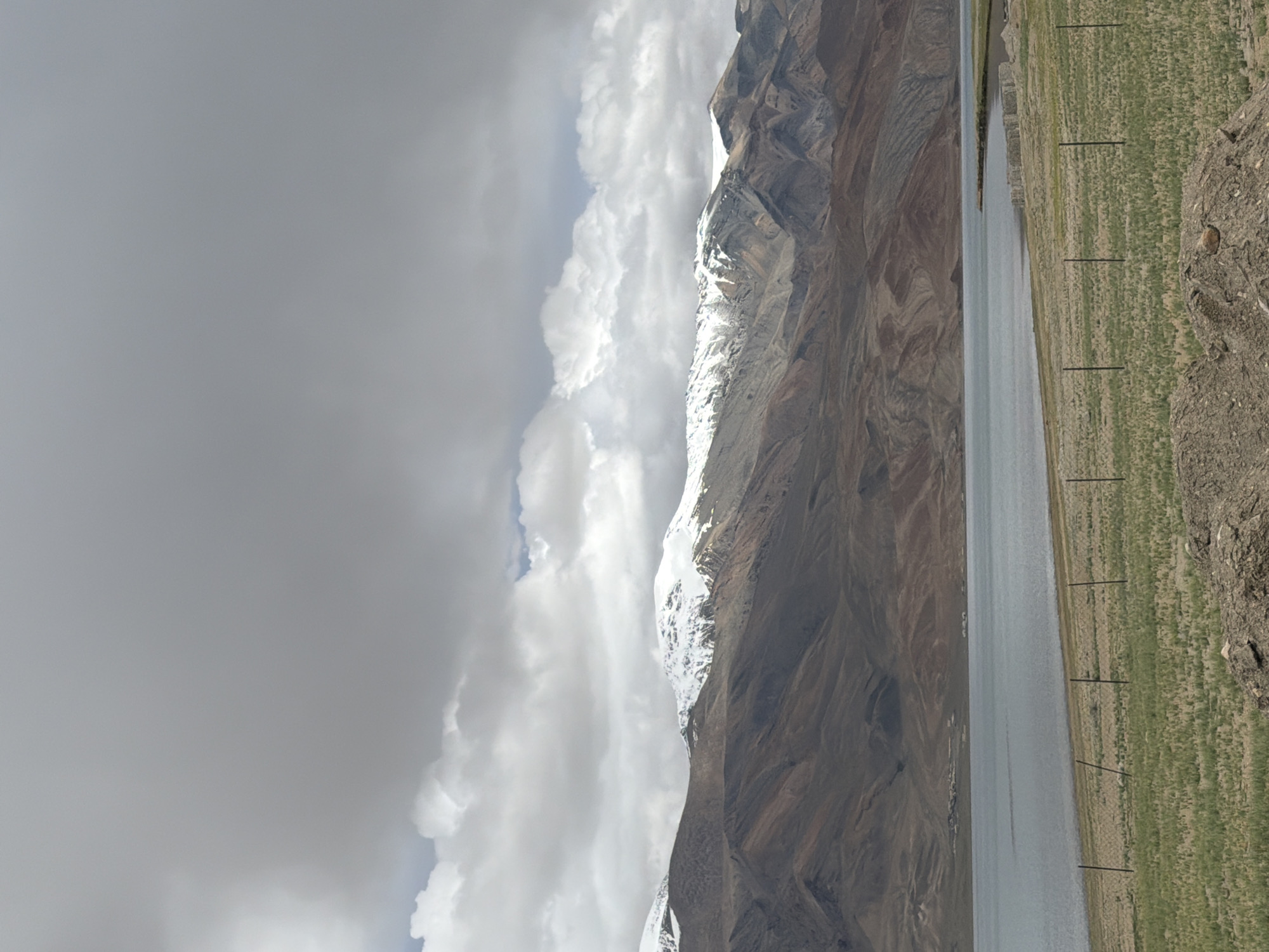 An overcast sky looms above a vast, serene lake in Karzok, India. Snow-capped mountains with rugged, brown slopes form a dramatic backdrop to the tranquil water. In the foreground, a grassy field stretches towards the lake, punctuated by a few thin, dark fence posts. The scene is natural and undeveloped, suggesting a remote and untouched landscape.