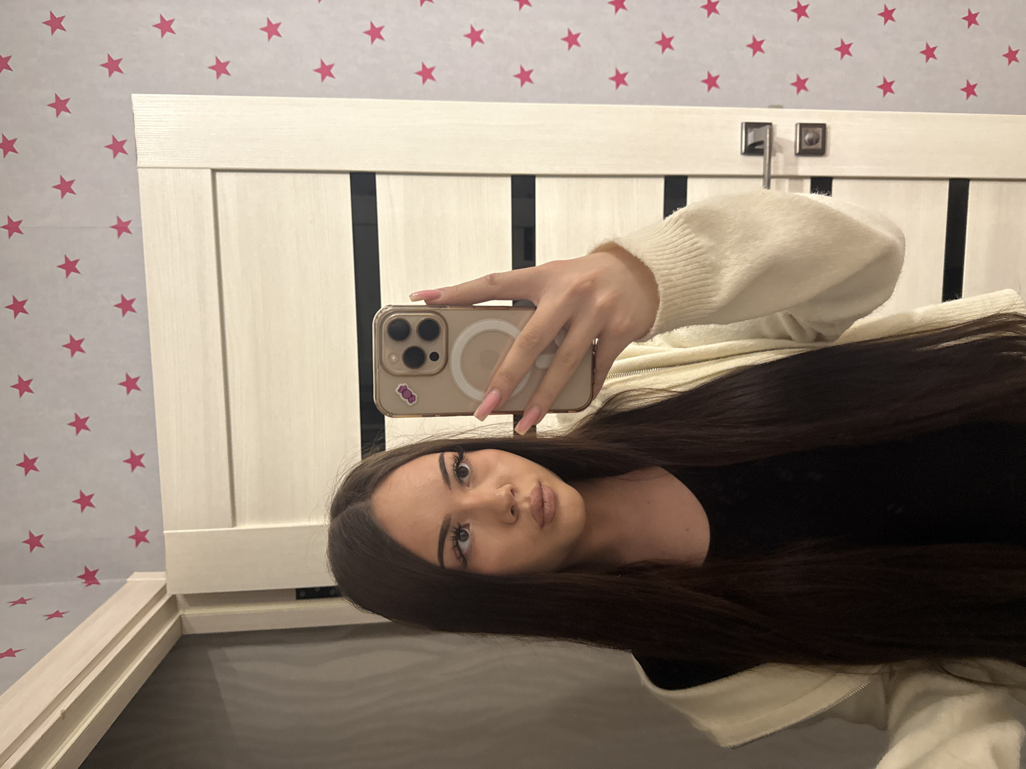 The image captures a young woman taking a selfie indoors, likely in a bedroom in Naberezhnye Chelny, Russia. She has long, dark brown hair, dark eyes, and a neutral expression, looking directly at the camera. Her subtle makeup includes winged eyeliner and nude lipstick. She wears a dark top under a cream-colored knitted cardigan.
In her right hand, she holds a light-colored smartphone, which appears to be an iPhone, featuring a clear or light-toned case with a visible circular MagSafe accessory and a small purple sticker. Her nails are long and painted a light pink.
The background consists of a light-colored wooden headboard or bed frame with vertical paneling, topped by two dark, square-shaped metallic accents. To the left, the wall is adorned with wallpaper displaying a pattern of scattered pink stars on a light background. The lighting is consistent with an indoor setting. No discernible text is clearly visible on the phone sticker.