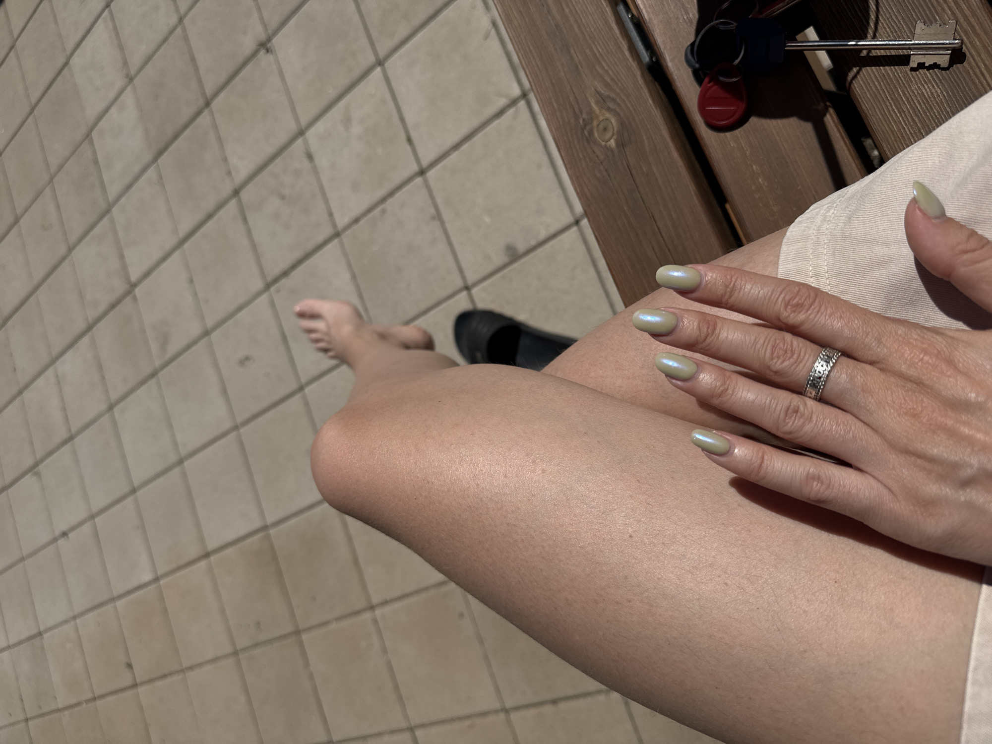 A close-up shot focuses on a person's legs and hands as they sit on a wooden bench outdoors. The person's left leg is bent at the knee, with a black sandal visible on their foot. Their right hand is in the foreground, displaying a silver ring and light green nail polish. A set of keys with red and black tags rests on the bench next to the person's legs. The background consists of a tiled patio area and a portion of the wooden bench. The lighting suggests it is a sunny day. There is no information available to identify the location within the city of Krasnoyarsk, Russia.