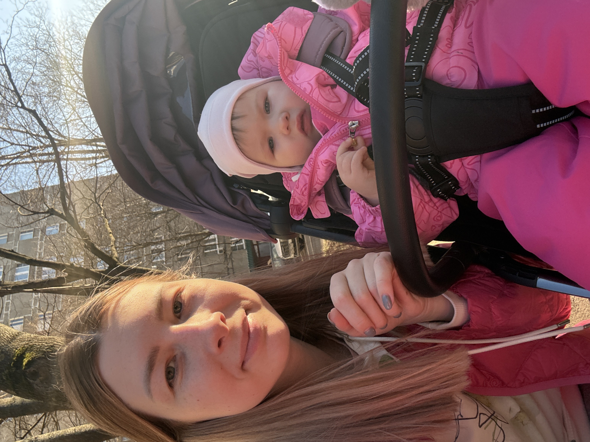 A woman with long blonde hair is taking a selfie with a baby in a stroller. The baby is wearing a pink jacket and a white hat. The woman is wearing a pink jacket. The photo was taken outdoors on a sunny day in what appears to be a city park. A building is visible in the background. Based on the architecture and the general appearance of the scene, it is possible that the location is in Moscow, Russia.