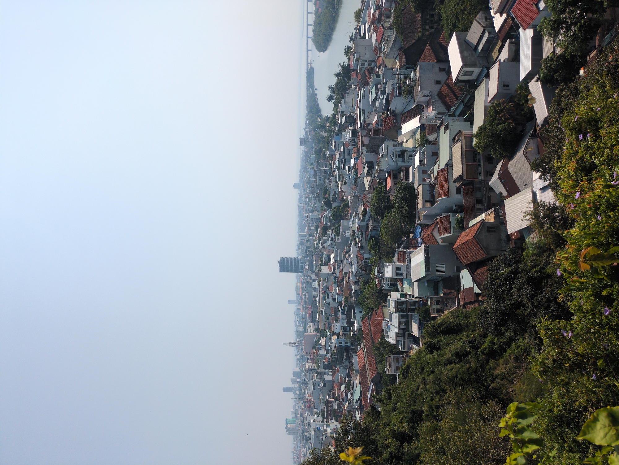 The image depicts an aerial view of a dense urban landscape in Tuy Hòa, Vietnam. The scene is dominated by closely packed buildings, primarily residential structures with varied roof colors, including red tile and light gray materials. A river runs through the city with a large bridge visible. Dense foliage and trees are on the right and bottom edges of the image, suggesting a hill or mountainous area. The air is hazy, reducing visibility towards the horizon. The city appears to stretch out along the river. A few taller buildings, including what looks like a skyscraper, interrupt the predominantly low-rise cityscape.