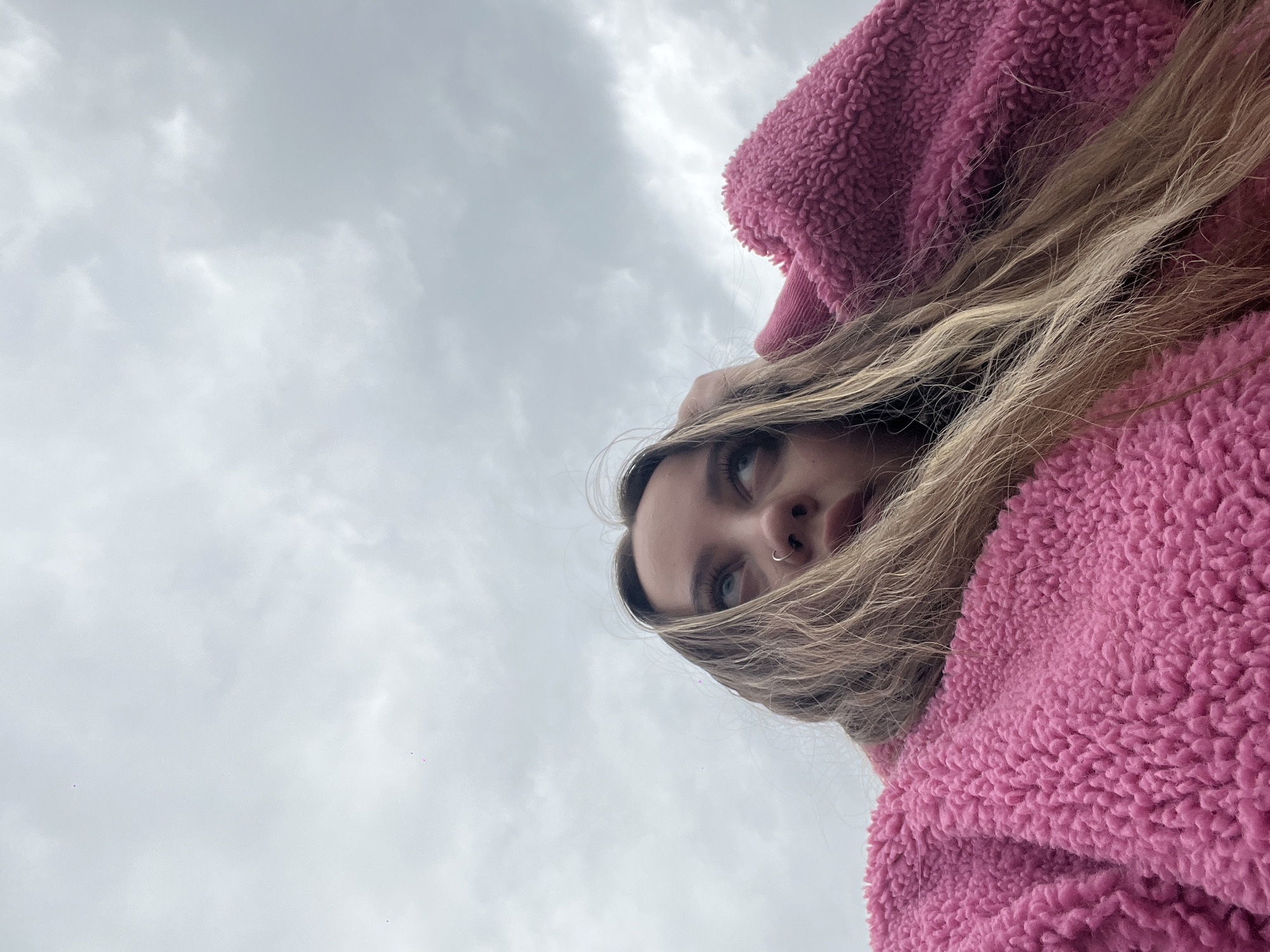 A low-angle shot captures a young woman with light brown hair wearing a pink, textured fleece jacket. She looks upward, and her face is visible in the upper right portion of the frame. Her eyes are blue, and she has a nose ring. The background consists of a grey, overcast sky. The photo's setting is unknown.