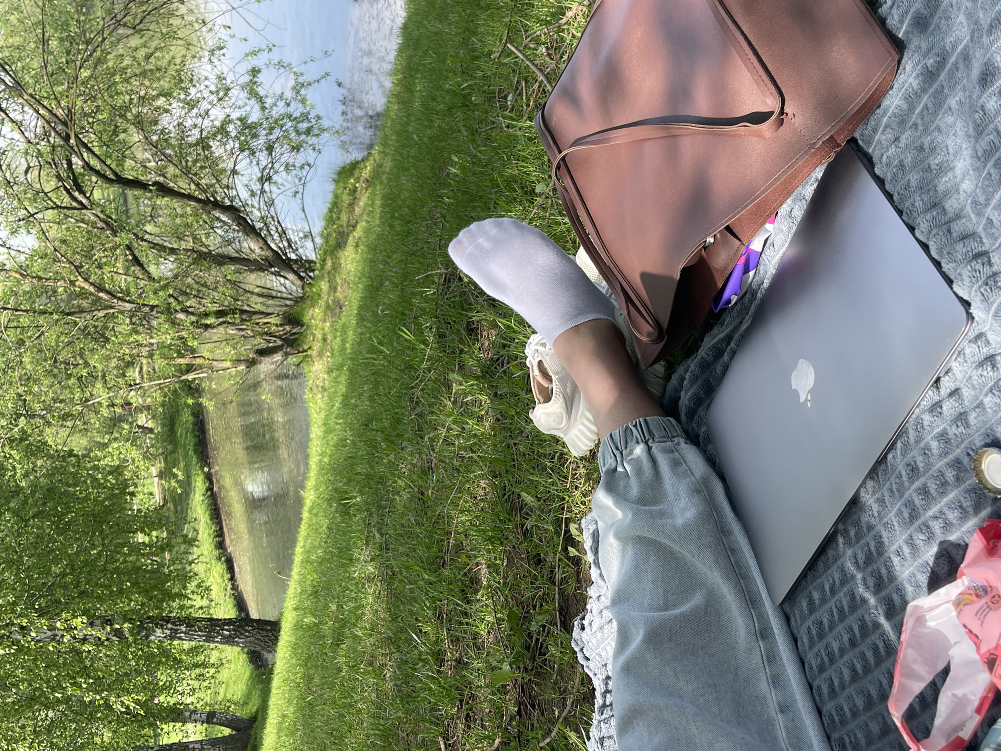 The image shows a person sitting on a gray textured blanket in a grassy area next to a body of water. The body of water is possibly a river or pond, judging by the vegetation and the landscape. The person is wearing light blue jeans and has one foot with a white sock and shoe visible. Next to their leg is a silver laptop and a brown leather bag. There is a bag with pink packaging to the right of the laptop. The environment appears to be a park or recreational area with trees lining the edge of the water. This image cannot be identified as being in Moscow, Russia.