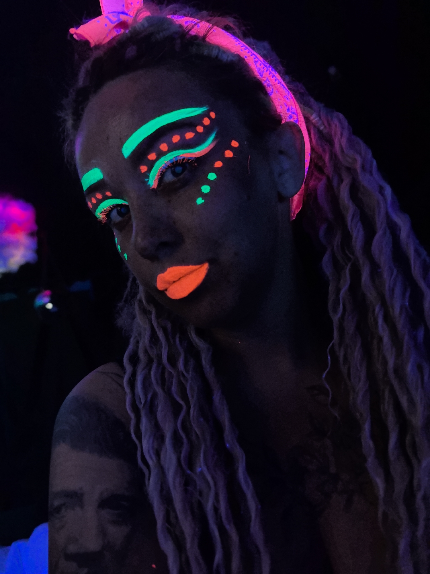 A close-up photo depicts a person with light skin and long, blonde dreadlocks under blacklight. The person is adorned with neon green and orange makeup, including a green stripe above each eyebrow, orange dots under each eye, and orange lipstick. A pink bandana is tied around their head. A tattoo of a man's face is visible on their upper left arm. The background is dark, with a faint, blurry pink and purple light source to the left. The photo's location is unknown and there is no information indicating a location in Noviy, Russia.