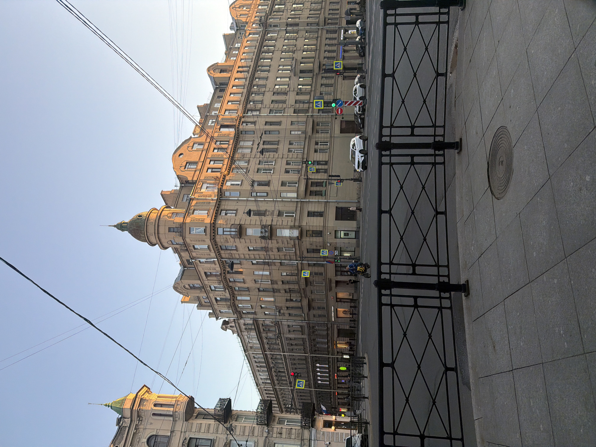 The image depicts a wide street intersection in Saint Petersburg, Russia, in the late afternoon or early evening, as evidenced by the warm, directional sunlight illuminating the facades of the buildings. The architecture is characteristic of the city, with ornate, multi-story buildings featuring mansard roofs, bay windows, and classical detailing.
In the foreground, a grey tiled sidewalk is bordered by a black metal railing with a diamond pattern. Beyond the railing, a two-lane road is visible, with several cars driving or stopped. Traffic lights are present at the intersection, some showing green signals. Pedestrian crossing signs are also visible.
The main focal point is a large, cream-colored building that dominates the right side of the frame. It has a rounded corner section topped by a green dome and spire. Another building to the left, also cream-colored, has a distinctive bay window with a green pointed roof.
Several pedestrians are present, including a person walking on the sidewalk on the left, a couple of people near the shops on the ground floor of the main building, and two individuals on a yellow scooter driving down the street. Overhead, power lines crisscross the sky, a common sight in urban environments. The sky is clear and blue. The scene conveys a sense of a bustling but orderly city intersection.