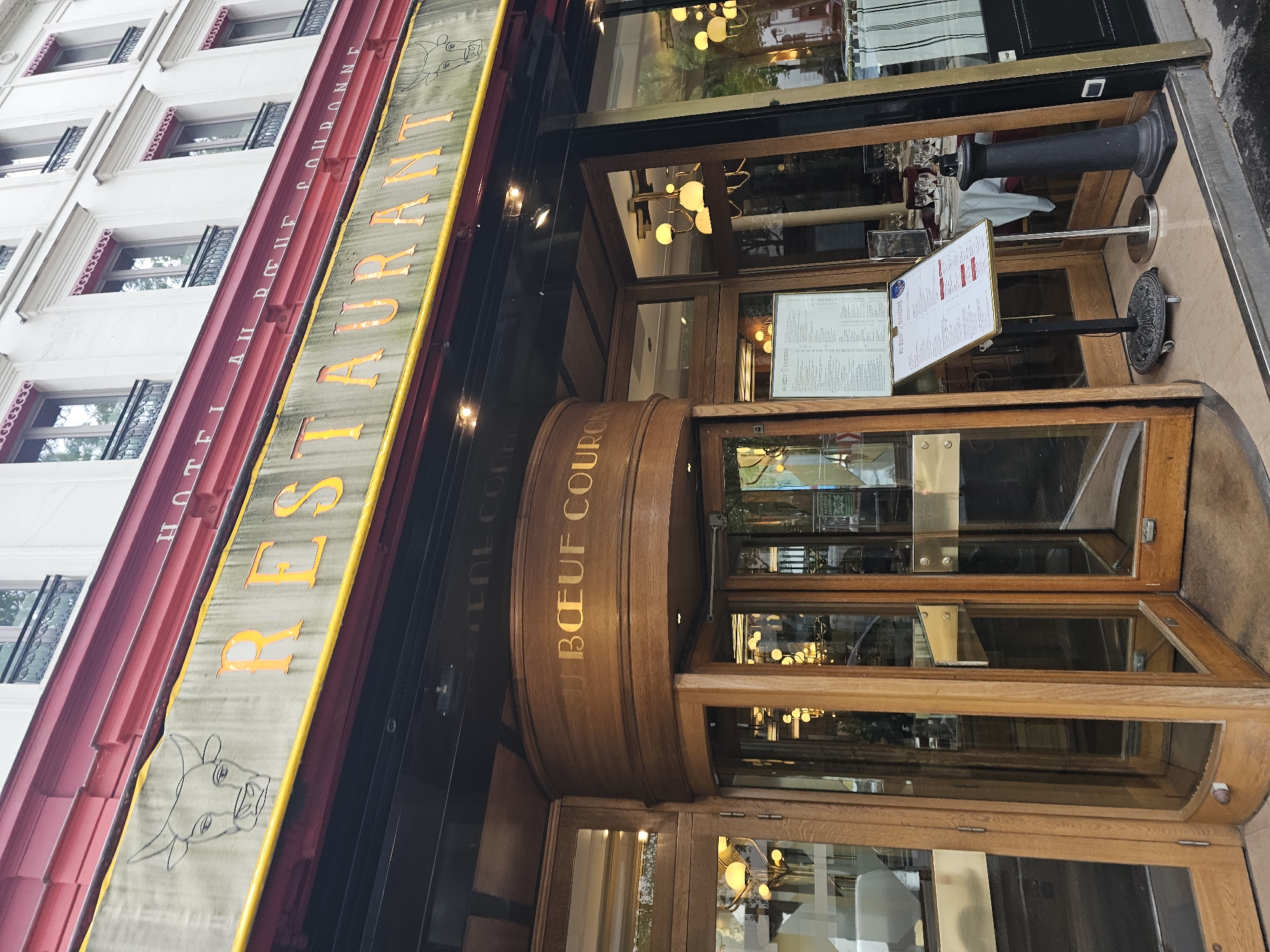The image captures the exterior of "Restaurant Au Boeuf Couronne," a restaurant located in Paris, France. The building has a red facade with white windows. A long, yellow awning with the restaurant's name in orange lettering hangs above the entrance. A cow's head is sketched on the left side of the awning. Below the awning, the restaurant's name is also carved in gold lettering on a wooden, semicircular structure that extends over the entrance. The entrance features revolving wooden doors. A menu on a stand sits outside the entrance. The surrounding environment is an urban street.