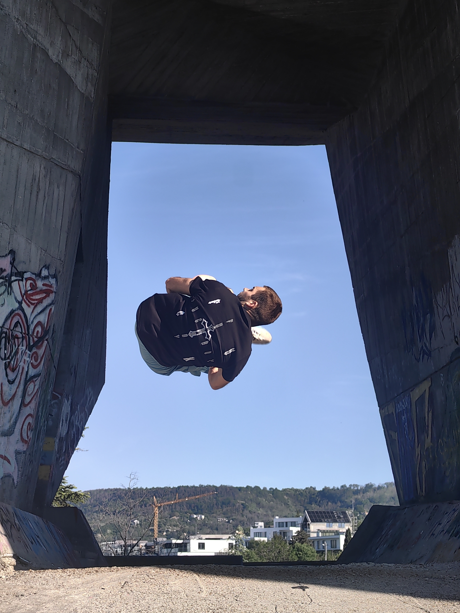 A man is performing a backflip in mid-air within a concrete structure in Varna, Bulgaria. The structure appears to be a bridge or tunnel entrance, with graffiti visible on the concrete walls. The man is wearing a black t-shirt with a white design, green shorts, and white shoes. In the background, there are buildings, trees, and a hill. A construction crane is also visible. The sky is clear and blue. The ground is covered with gravel.