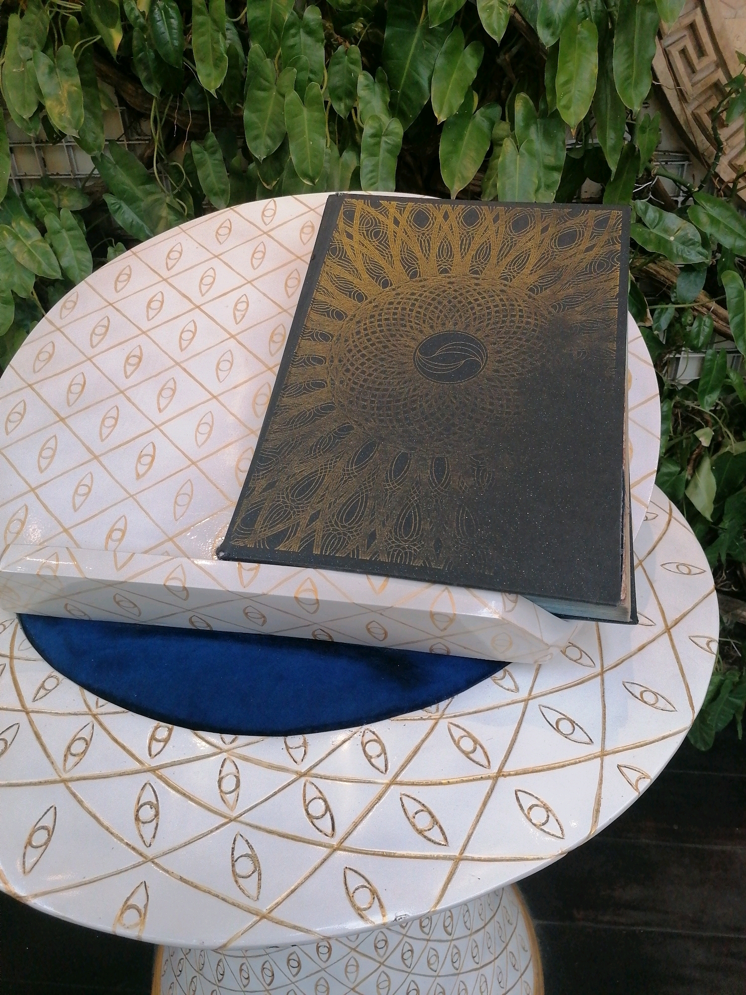 A close-up, overhead shot captures a black, hardcover book with a gold-patterned cover, resting on a white, tiered table with a diamond-shaped pattern of gold eye-like symbols. The book's cover features an intricate, mandala-like gold design with a central oval emblem. The table's surface is smooth and reflective, with a slight sheen. The background consists of lush, green foliage and a hint of textured wall. The image appears to be staged, likely for a product shot or artistic presentation, and shows no signs of movement or human interaction. The scene is well-lit, highlighting the textures and details of the objects.