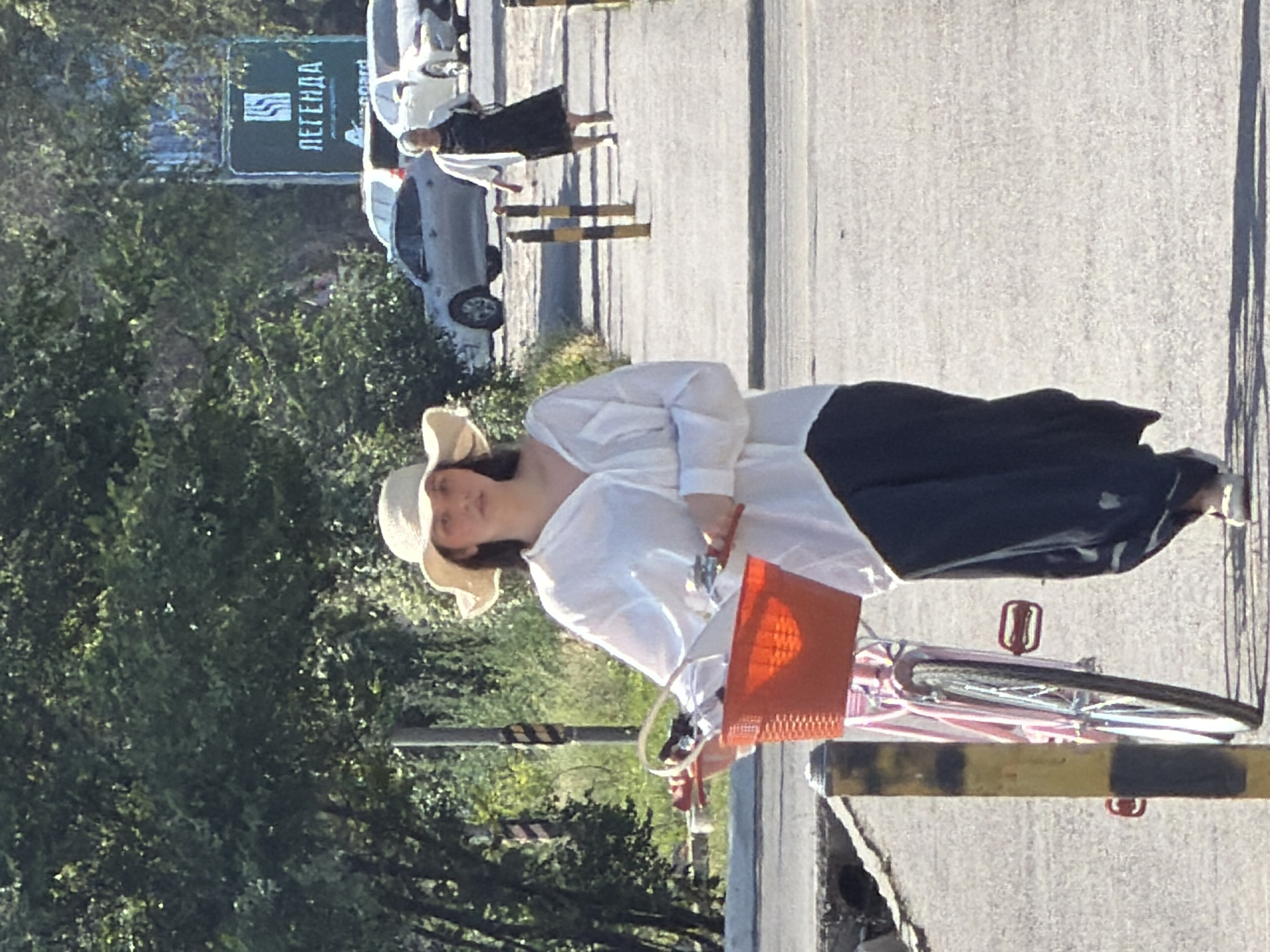 In an outdoor urban setting, a young woman wearing a wide-brimmed hat and a white oversized shirt walks with a pink bicycle. The bicycle has a bright orange basket attached to the handlebars, containing various items. She is wearing dark trousers and light-colored shoes. In the background, another woman in a black dress and white cardigan walks away from the camera. Several cars are parked and driving on a road behind them, and a green sign with white lettering, possibly an advertisement, is visible. The scene appears to be a sidewalk or pedestrian pathway adjacent to a road, with trees and foliage lining the left side of the image. The lighting suggests it is daytime.