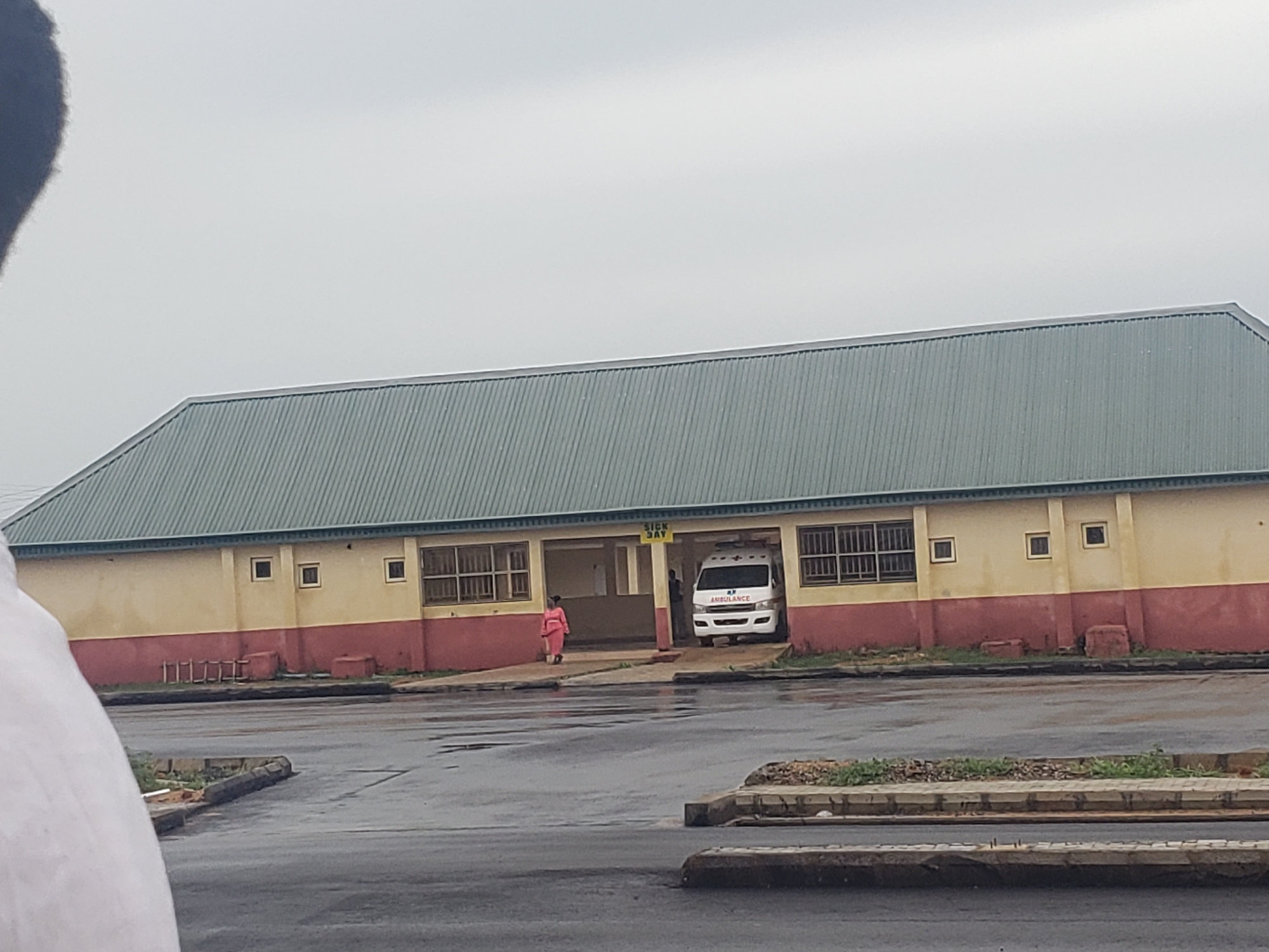 The image shows an outdoor scene in front of a building with a green, corrugated metal roof and yellow and red walls. A sign that reads "SICK BAY" is visible above an open entryway. An ambulance is parked inside the entryway. A person in a pink garment stands near the left side of the entryway, and another person is visible inside the entryway to the right of the ambulance. The ground in front of the building is wet and appears to be asphalt, with some patches of grass and debris. The sky is overcast. The image is taken from a low angle, with a portion of a person's white shirt or clothing visible in the foreground on the left.